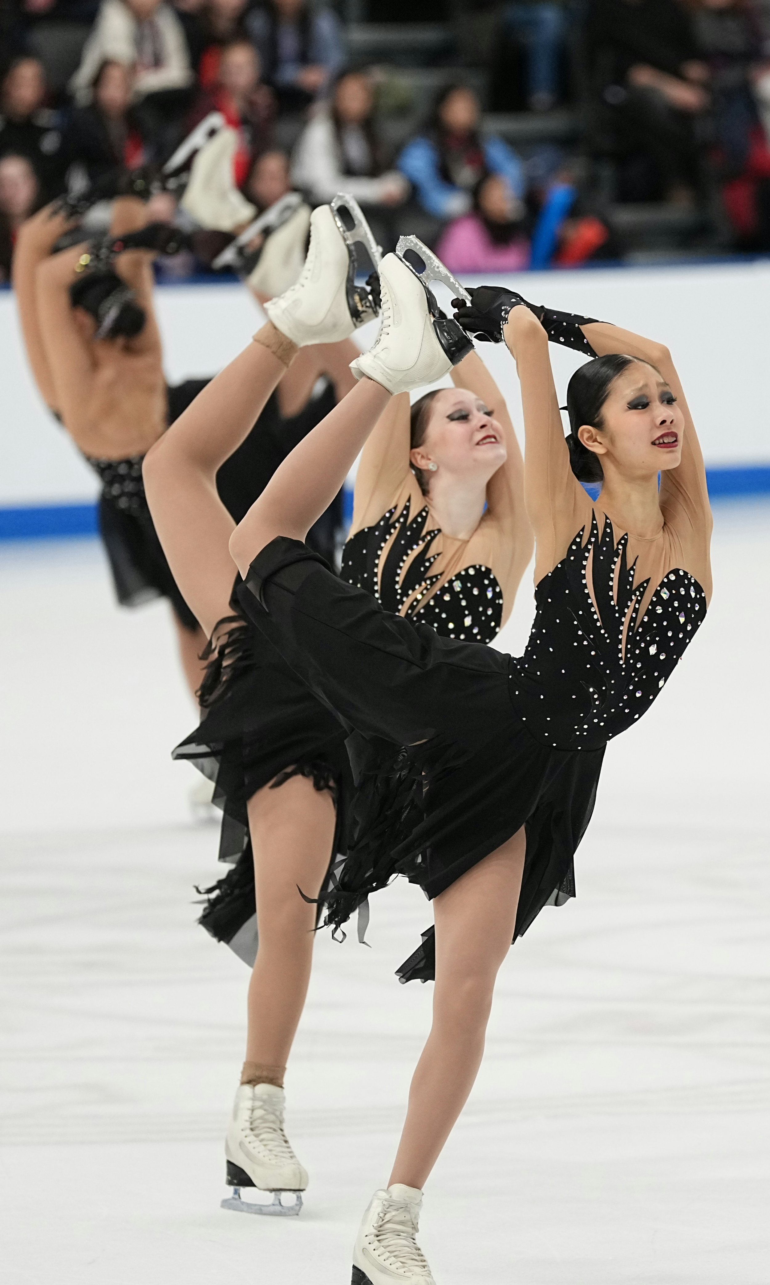 Figure skaters performing a synchronized routine on ice, dressed in black costumes with cut-out designs and embellishments, with an audience watching in the background.