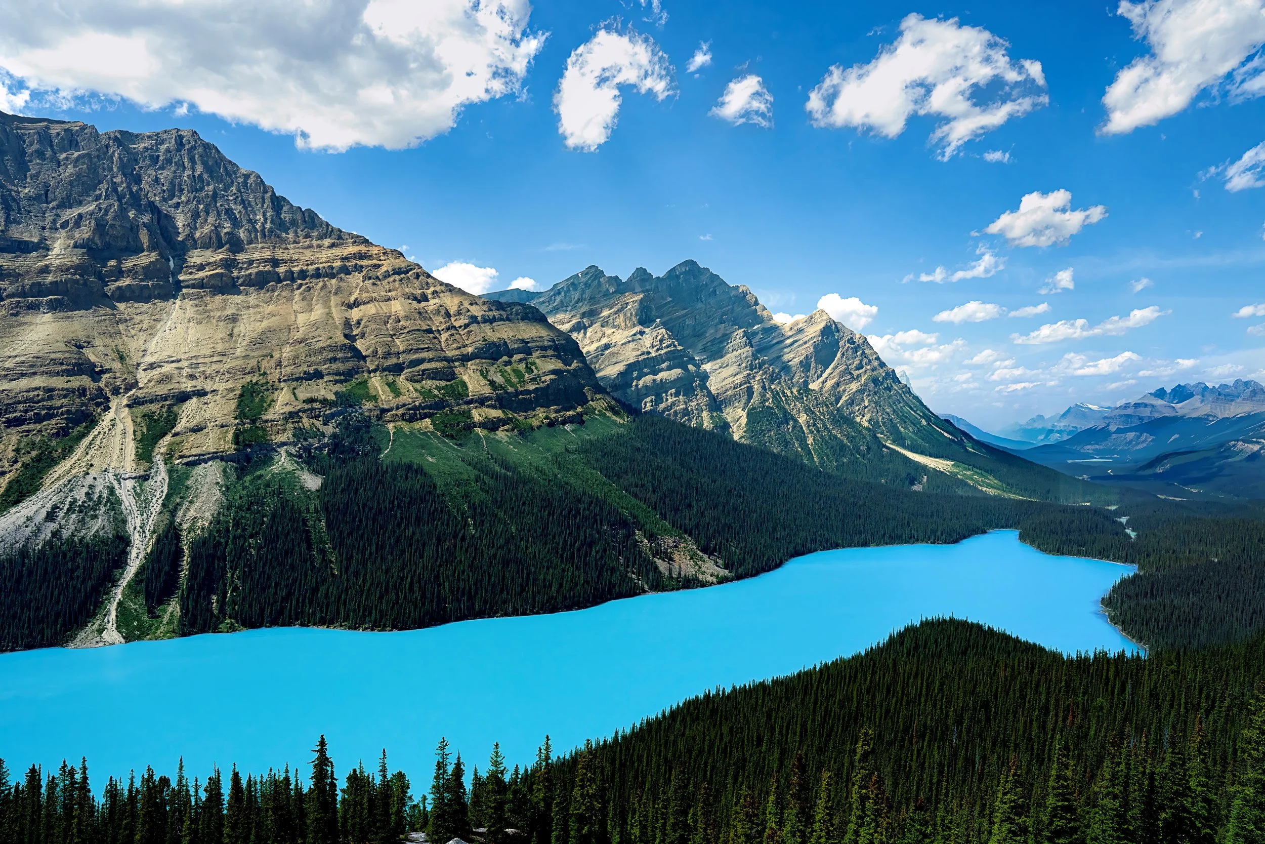 Scenic view of a bright blue lake surrounded by dense green pine forests and towering rocky mountains under a partly cloudy sky.