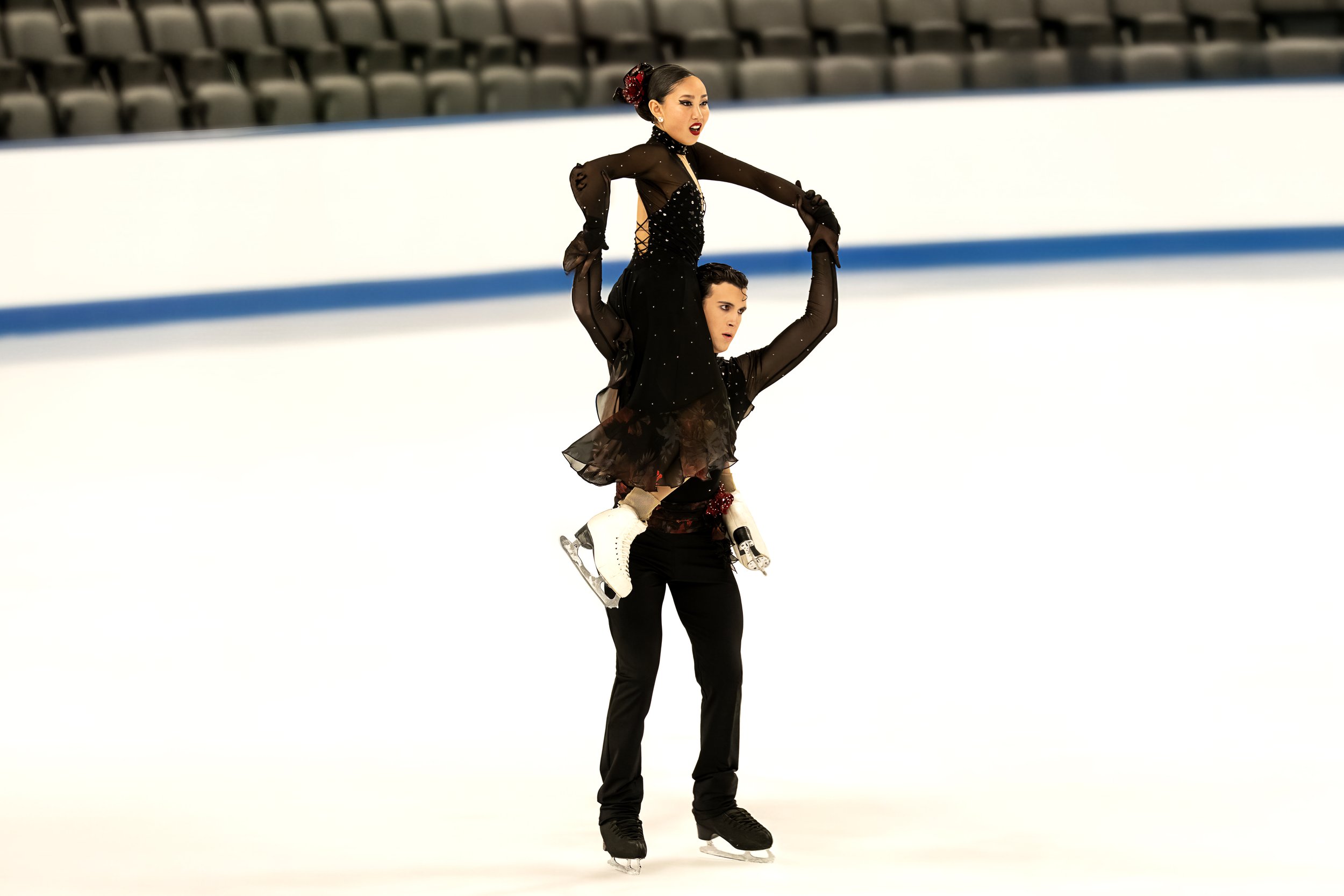 Two figure skaters performing a lift on the ice rink, with rows of empty seats in the background.