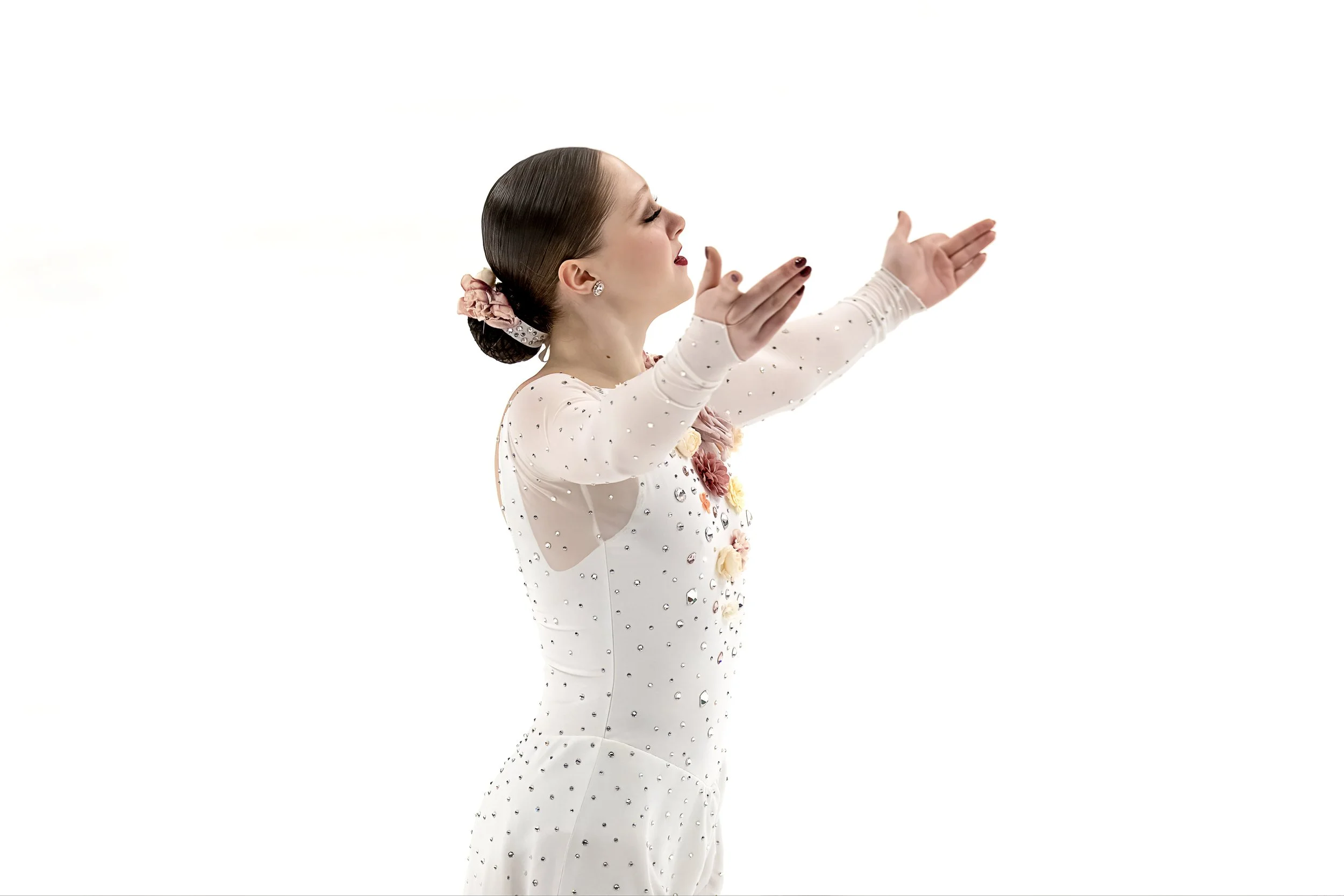 A woman with her hair in a bun, wearing a white dress with embellishments, appears to be figure skating or performing on ice, with her arms extended in a pose.