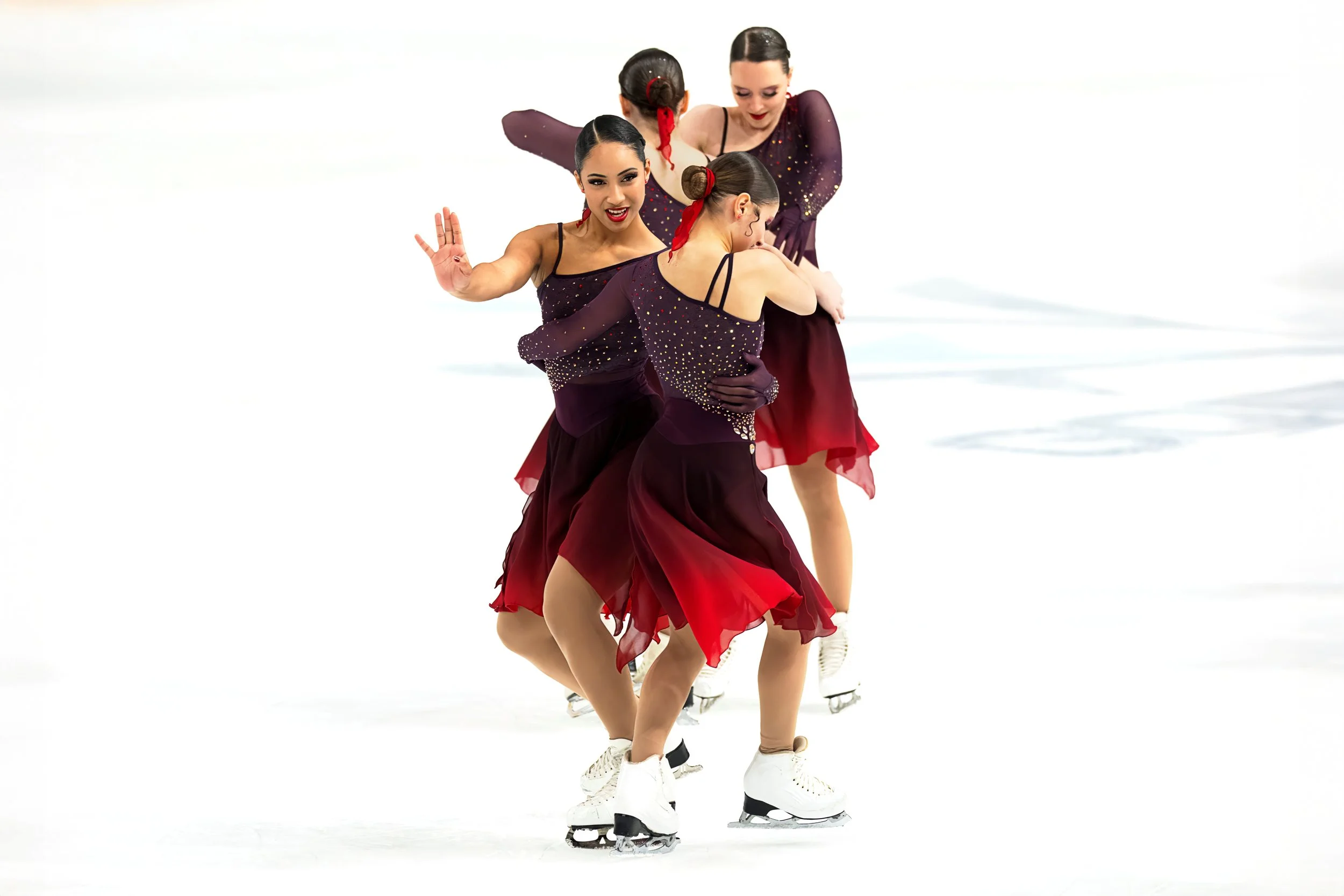 Four female figure skaters dressed in matching purple and red costumes performing a synchronized routine on ice skate with a white background.