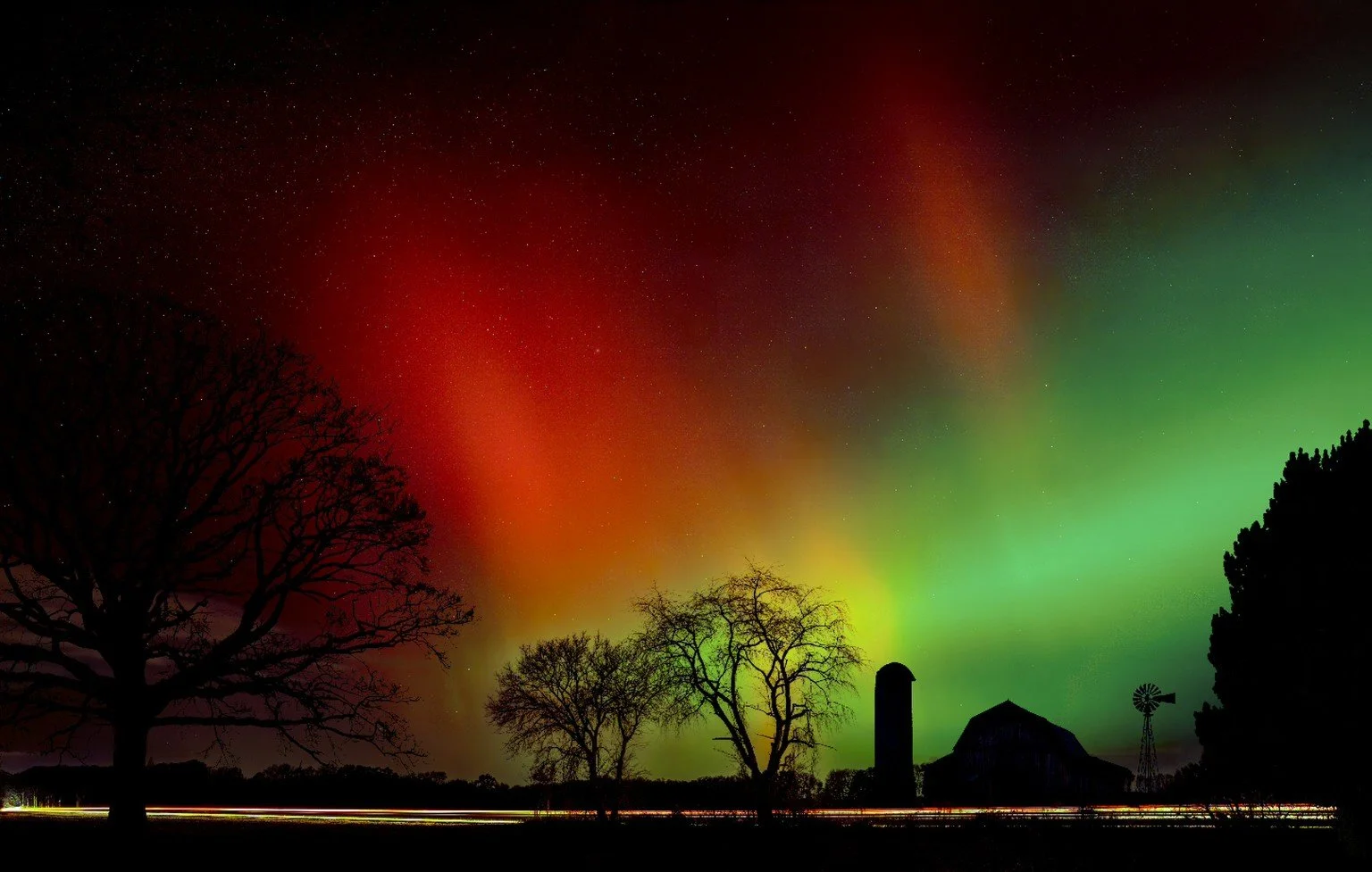 Nighttime landscape showing colorful northern lights in green, red, and orange across the sky, with silhouetted trees, a barn, a silo, and a windmill in the foreground.