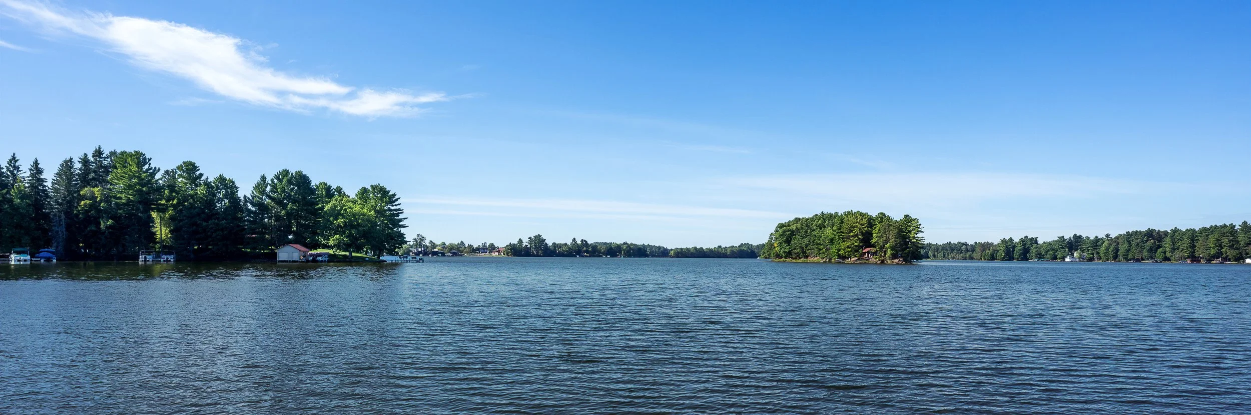 A scenic view of a lake with calm water, surrounded by green trees and small houses along the shoreline. The sky is blue with a few white clouds.