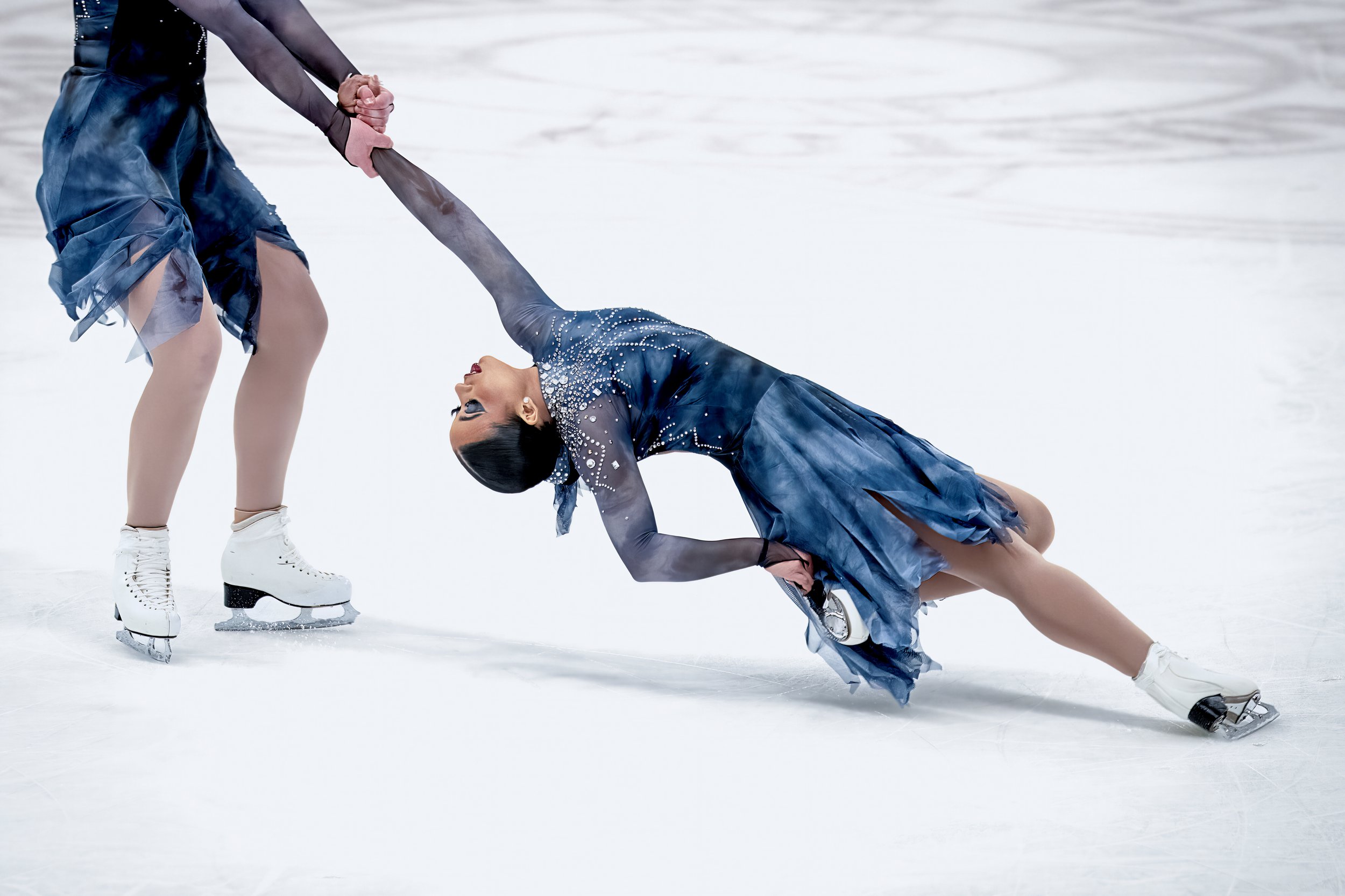 Figure skater performing a move on the ice, wearing a blue dress with rhinestones, with another person holding her hand on ice skates.