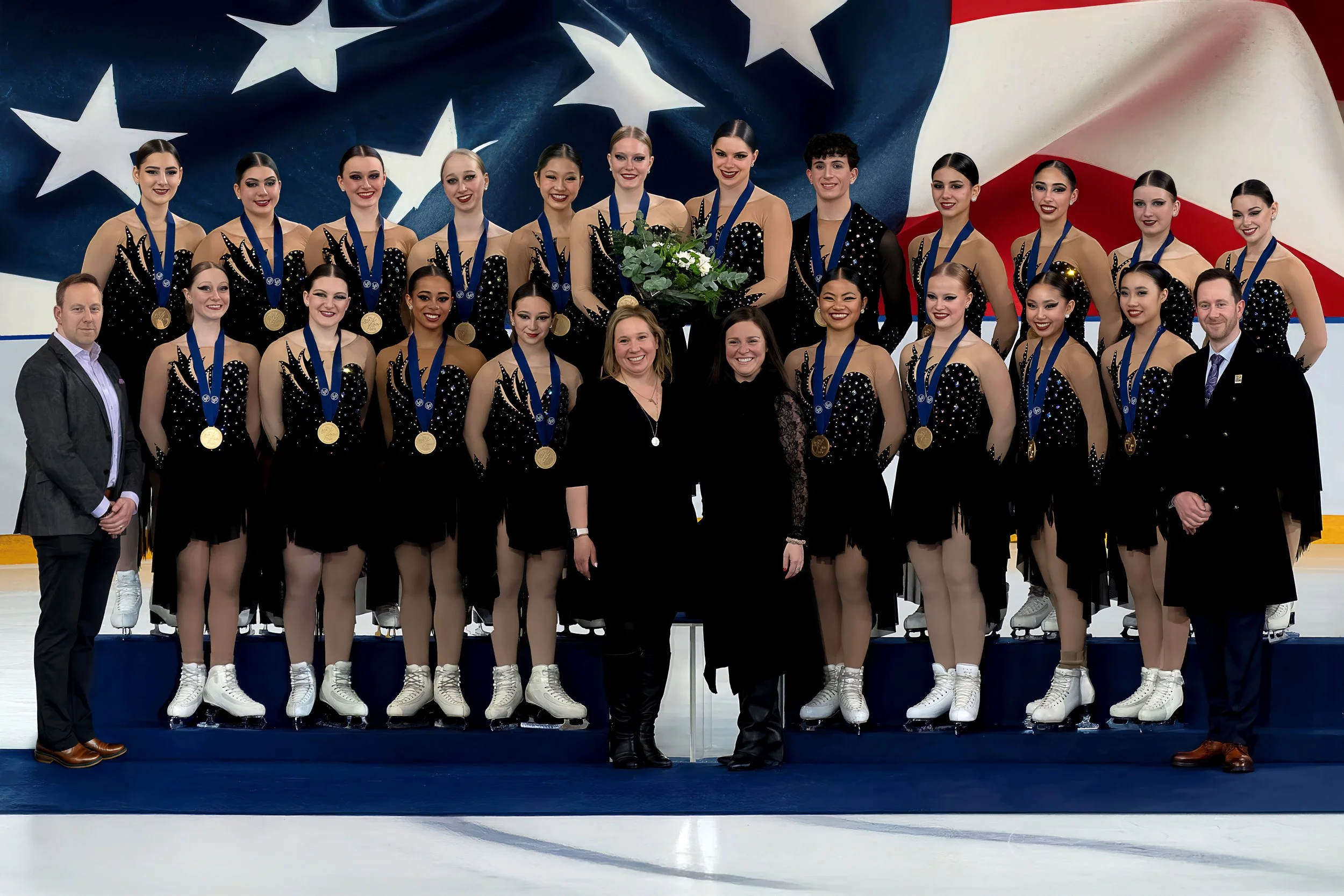 Group of synchronized figure skaters on the podium with medals and flowers, posing for a photo with coaches and officials in front of an American flag backdrop.