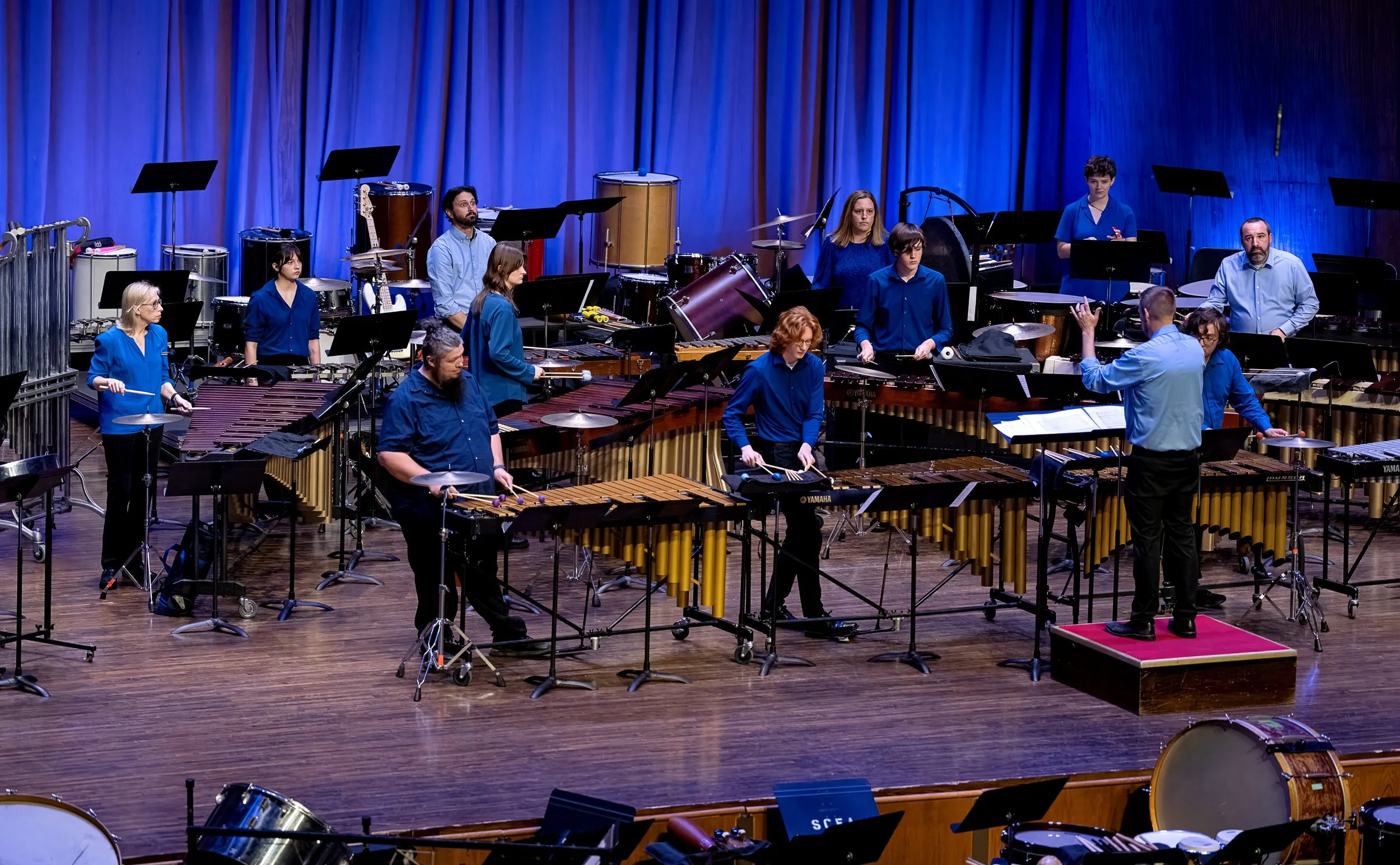 A group of musicians performing on stage, featuring various percussion instruments, with a conductor leading them.