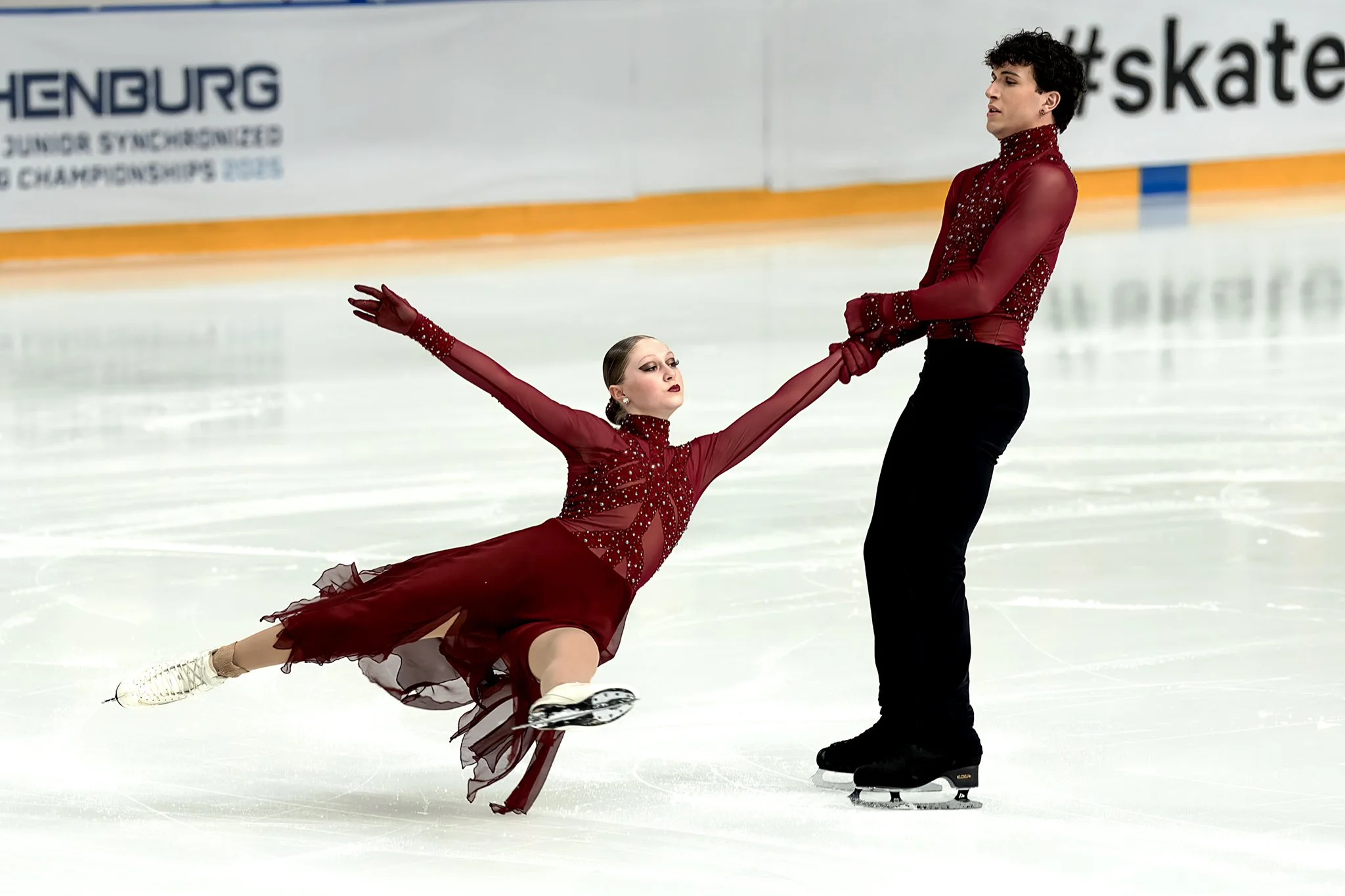 A pair of figure skaters performing on an ice rink, with one skater in a deep lunge position and the other standing, holding hands, both dressed in matching red costumes with sequins.