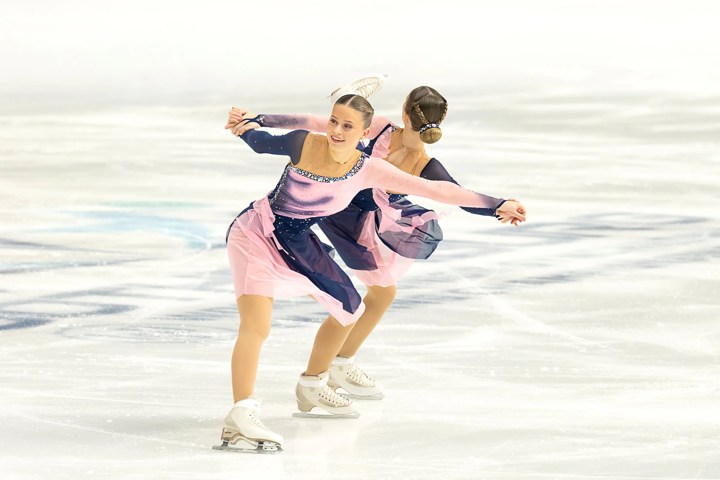 Two female figure skaters on the ice, performing a routine, dressed in colorful costumes with pink, navy, and beads, with a white background and ice surface.