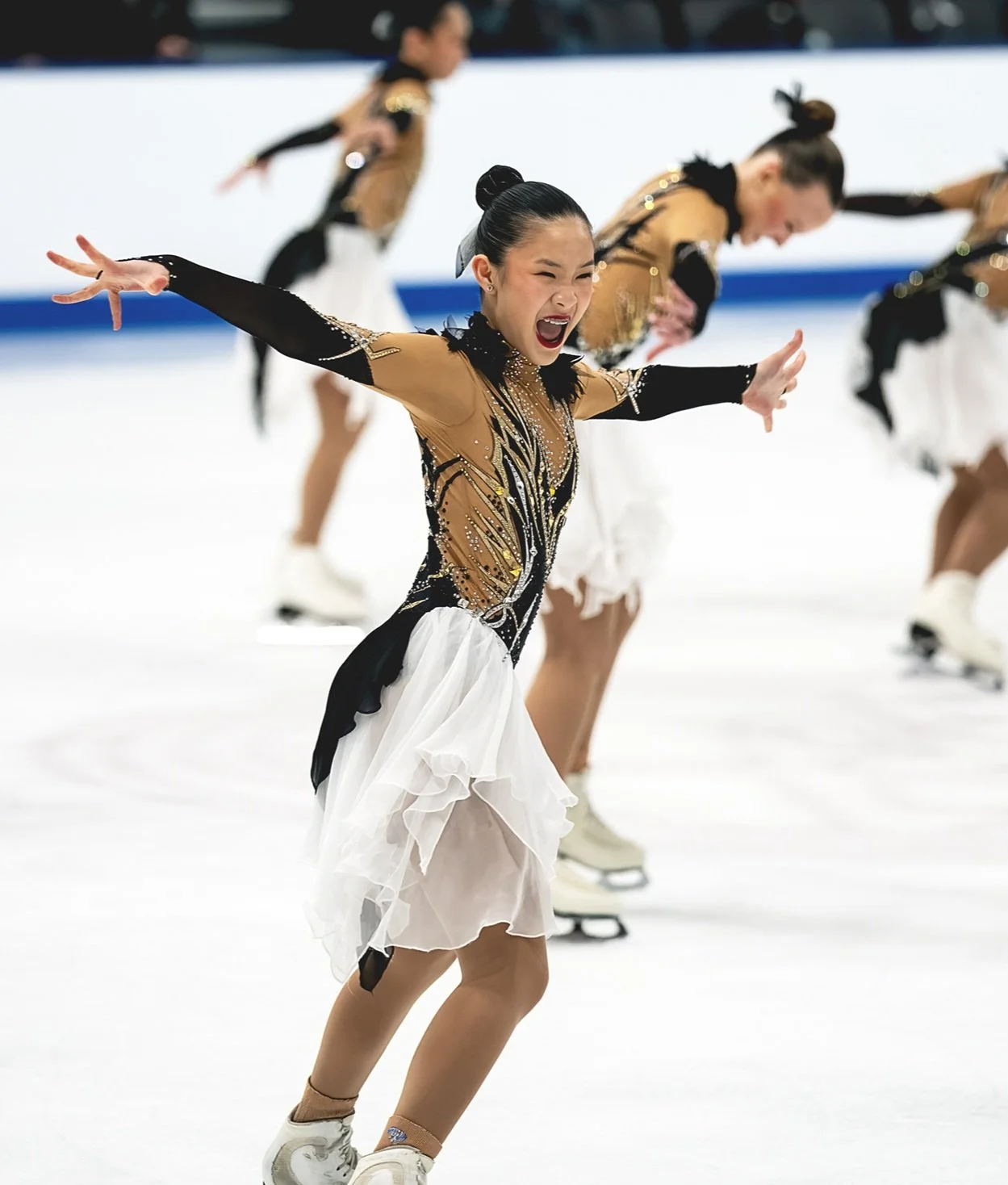 Figure skater performing on ice rink in a costume with black, gold, and white design, with other skaters in matching costumes in the background.