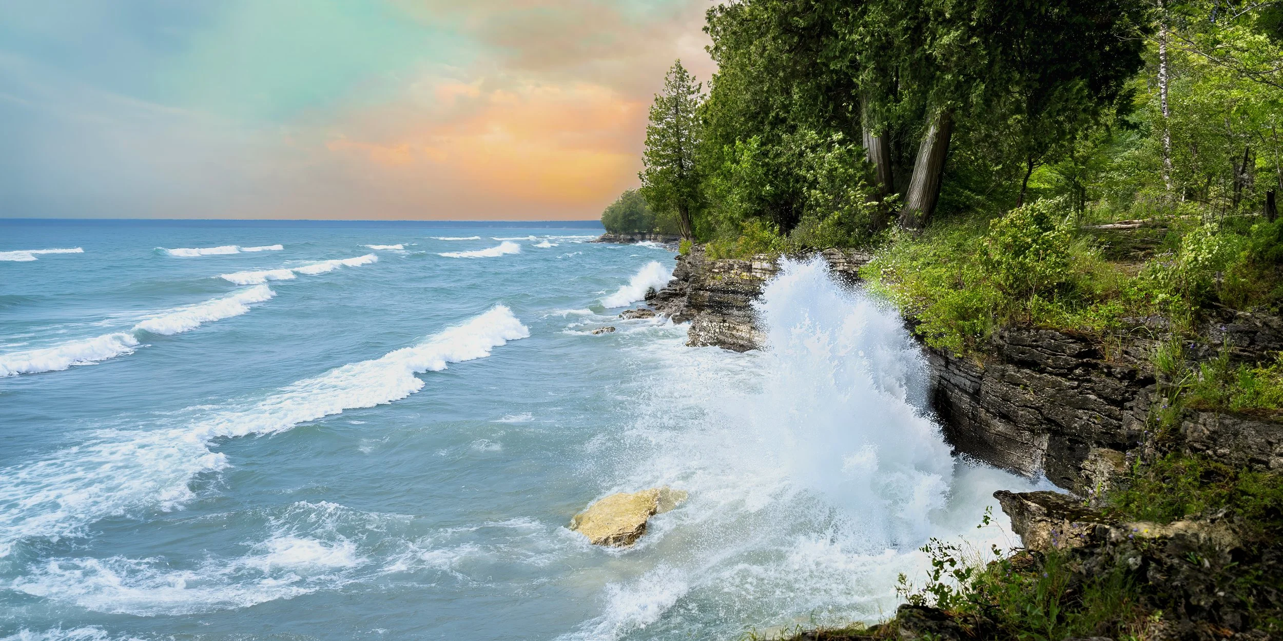 Waves crashing against a rocky shoreline lush with green trees, under a partly cloudy sky with a soft sunset glow.