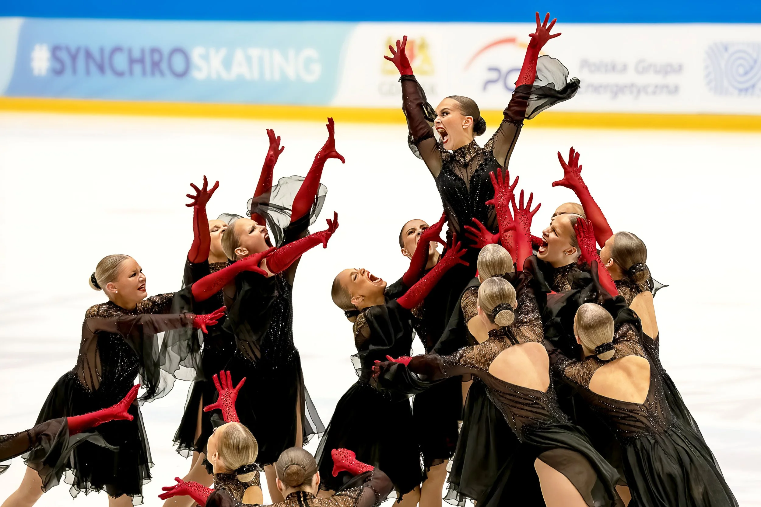 A synchronized skating team performing a routine on the ice, with one skater elevated and arms raised in a dynamic pose, all wearing coordinated black costumes with red gloves.