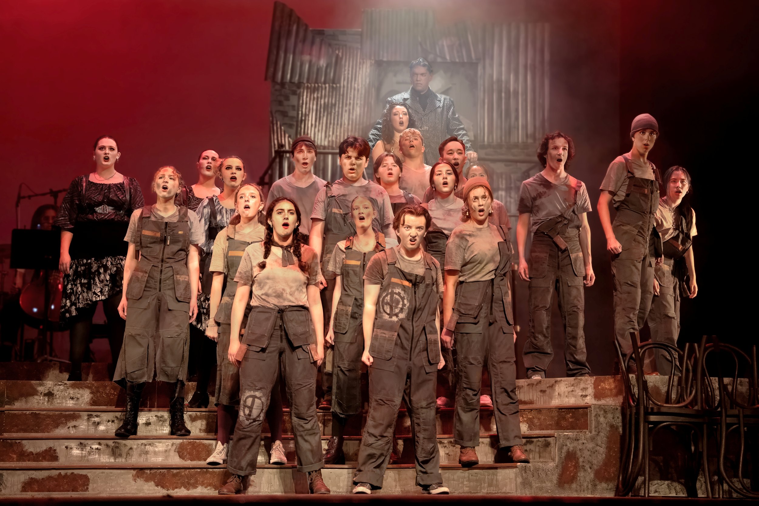 Stage performance of a group of young actors singing with serious expressions, dressed in distressed clothing and standing on stairs with a backdrop of a dilapidated building.