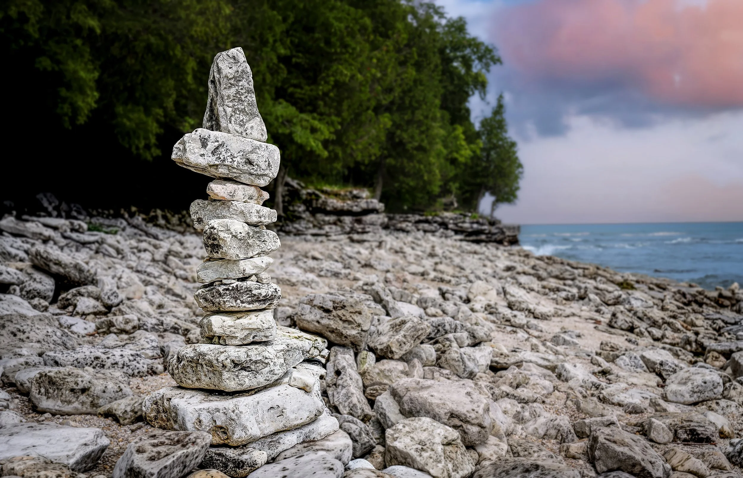 Stacked rocks on a rocky beach with green trees in the background and a cloudy sky at sunset.