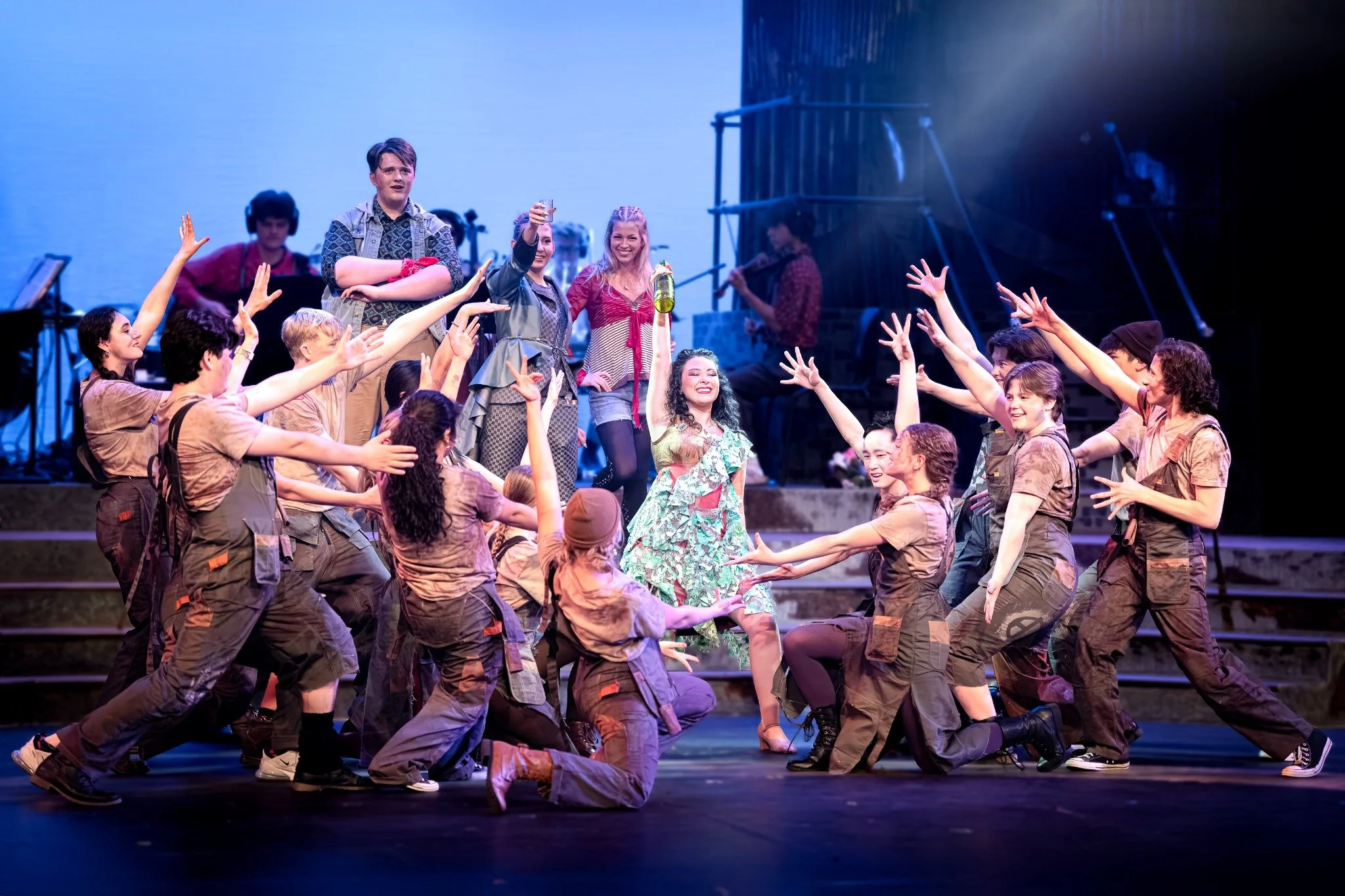 Theater scene with a woman in a floral dress kneeling in front, surrounded by dancers in brown outfits, some reaching arms out. In the background, people are standing with drinks, and musicians are on stage.