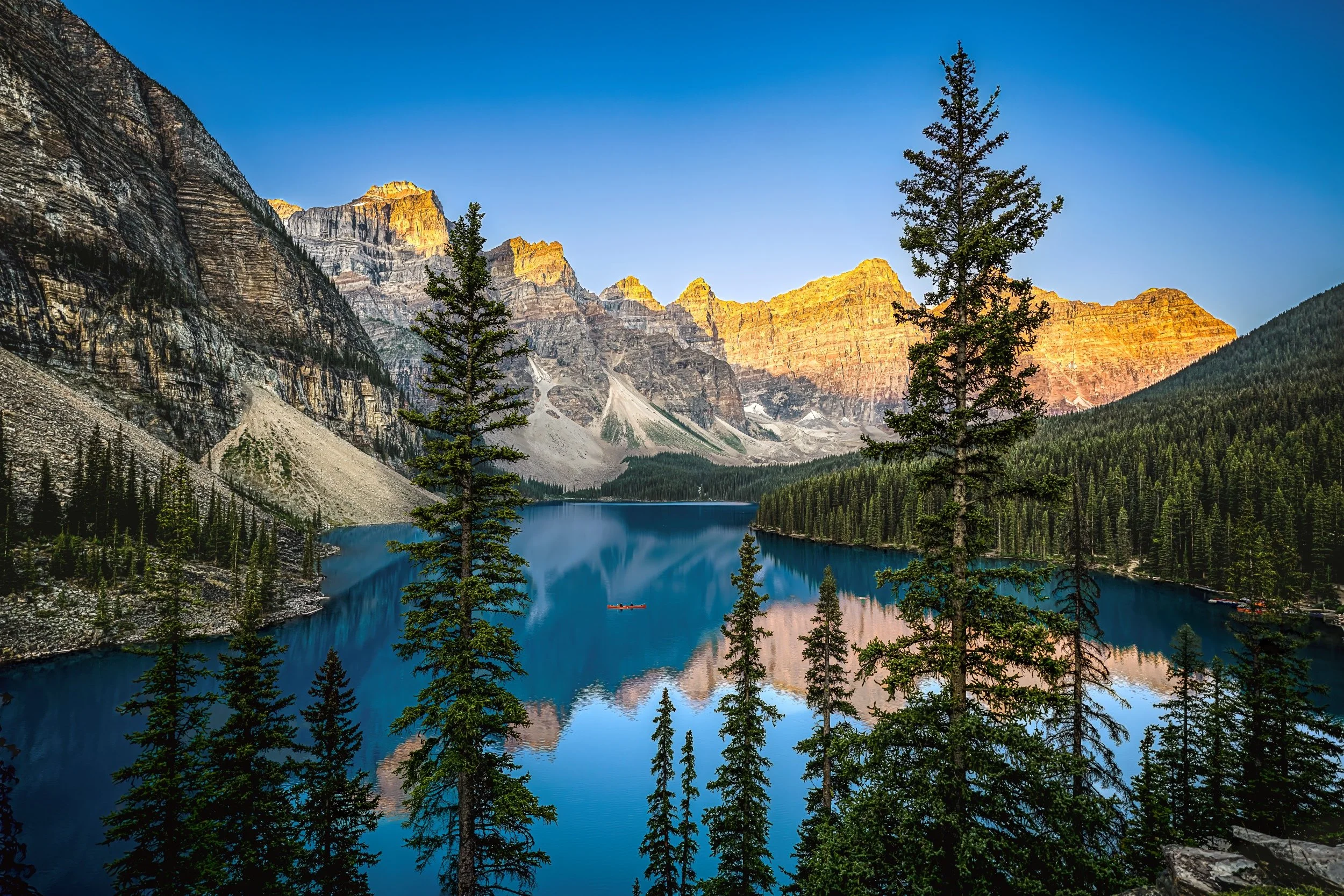 Scenic view of a mountain range at sunset with a reflective lake and pine trees in the foreground.