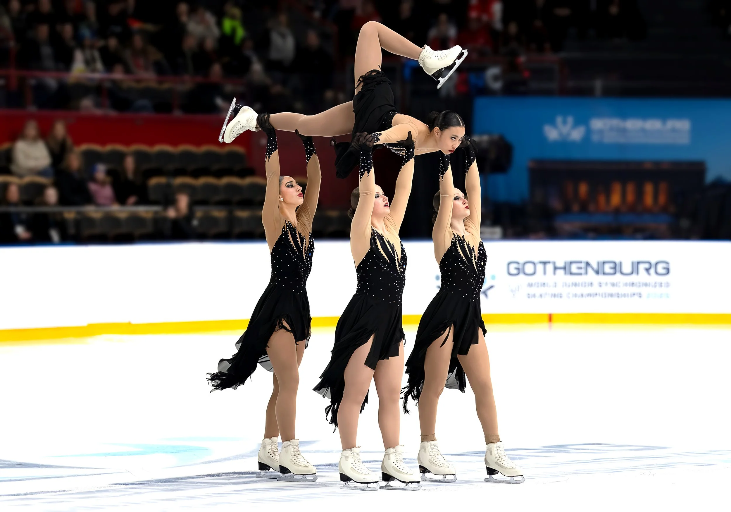 Six female ice skaters in black and beige costumes perform a synchronized move on an ice rink. One skater is lifted above the others, supported by five skaters holding her. The background shows an audience and a sign with partial text reading 'GOTHENBURG'.