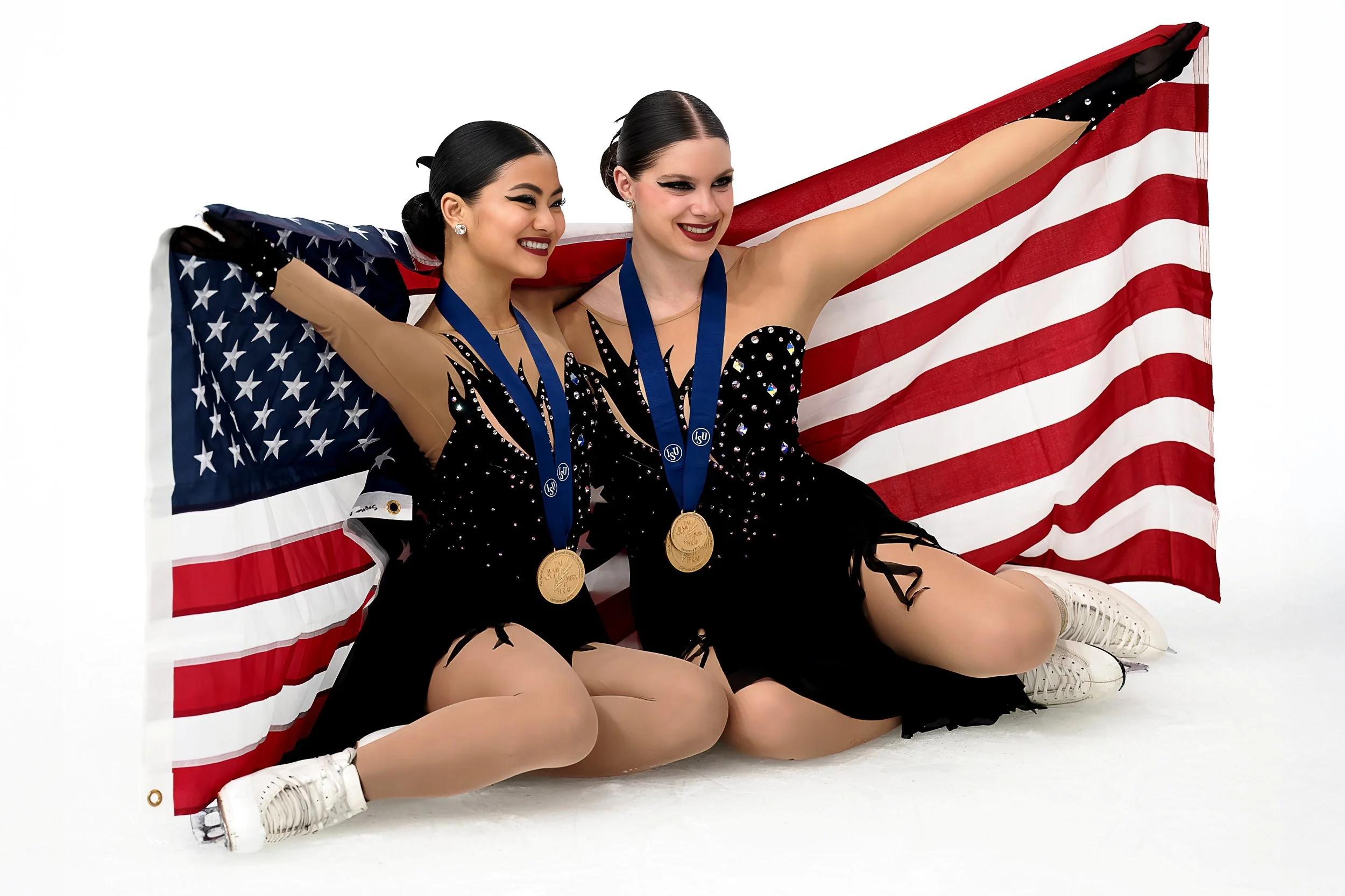 Two female ice skaters sitting on the ice, holding American flags over their shoulders, wearing black costumes with rhinestones, medals around their necks, and white figure skates, smiling after a competition.
