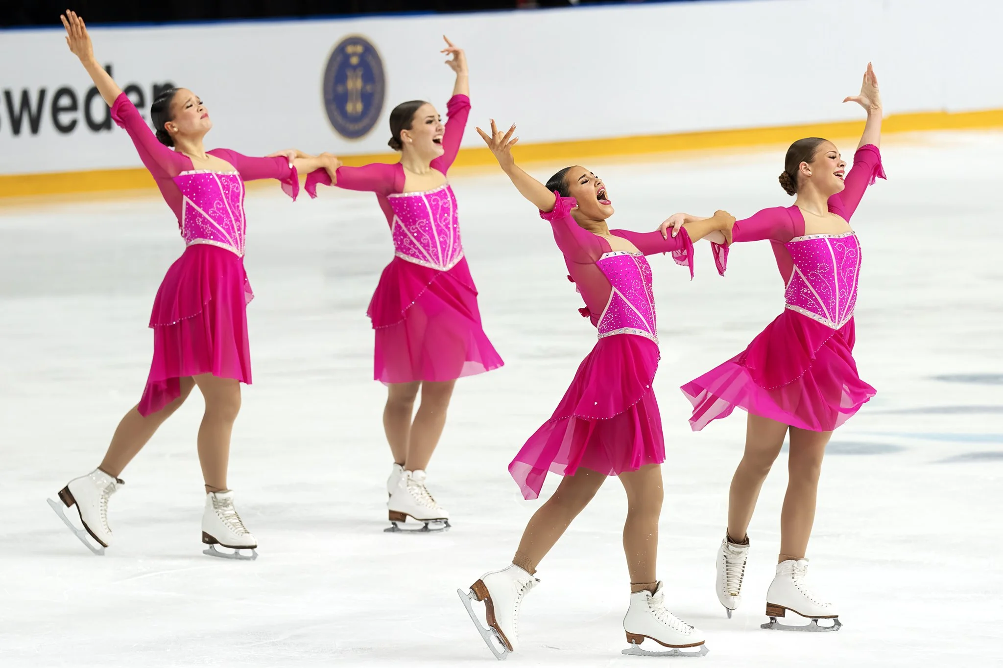 Four female figure skaters in matching pink costumes performing a synchronized routine on the ice rink.