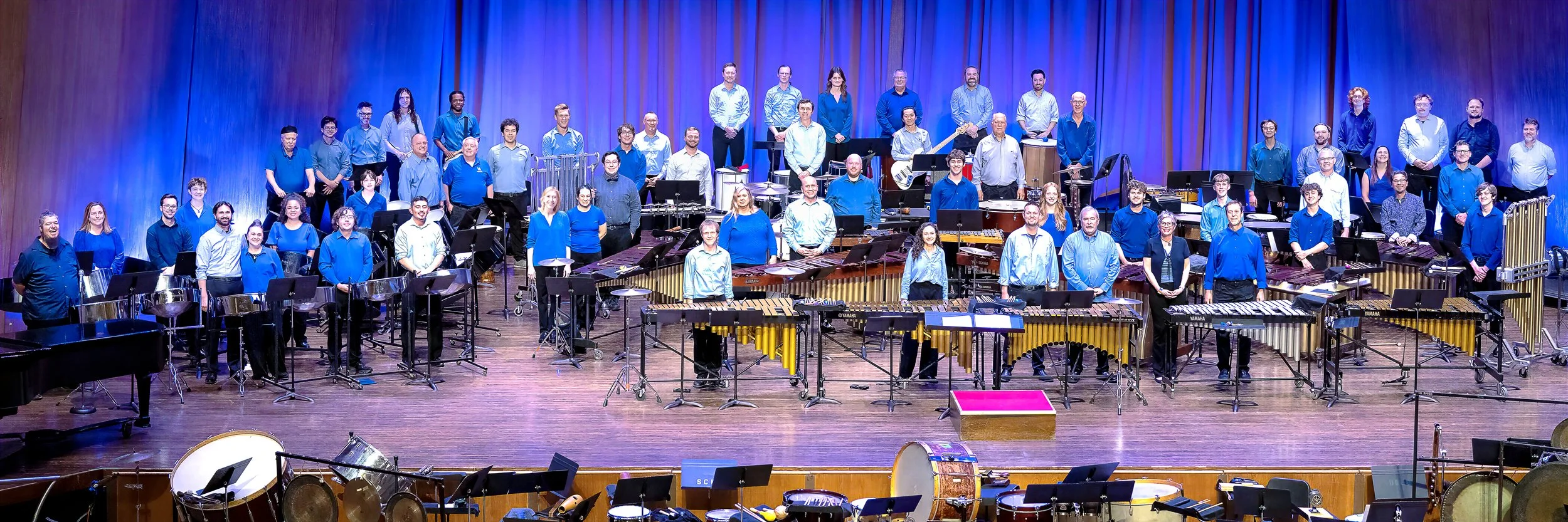 A large group of musicians, including percussionists, pianists, and wind instrument players, standing on a stage with blue curtains behind them, in a concert hall.