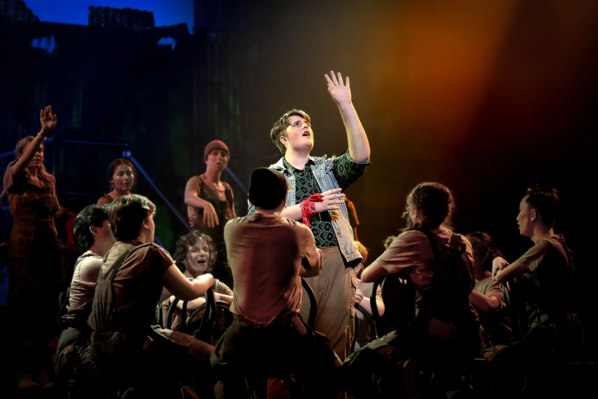 A group of people, mostly young adults, in a dimly lit setting with colorful stage lighting, with one person standing and reaching upward, while others sit or stand around in various poses.