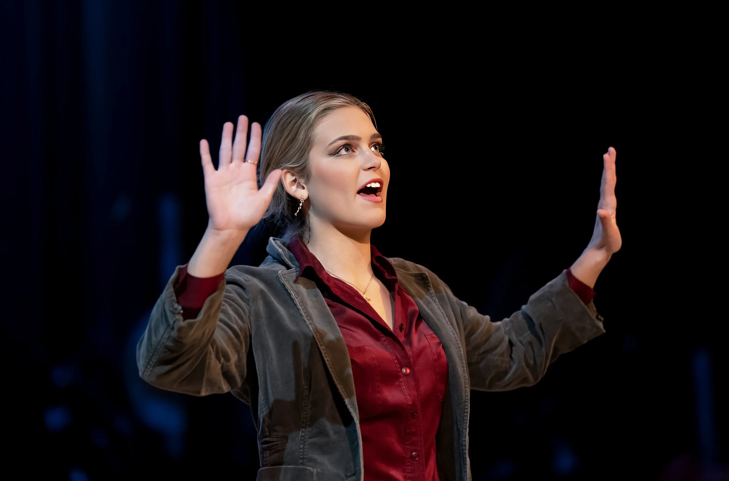 A woman with blonde hair, wearing earrings, a red shirt, and a gray jacket, appears to be speaking or explaining something with her hands raised, against a black background.