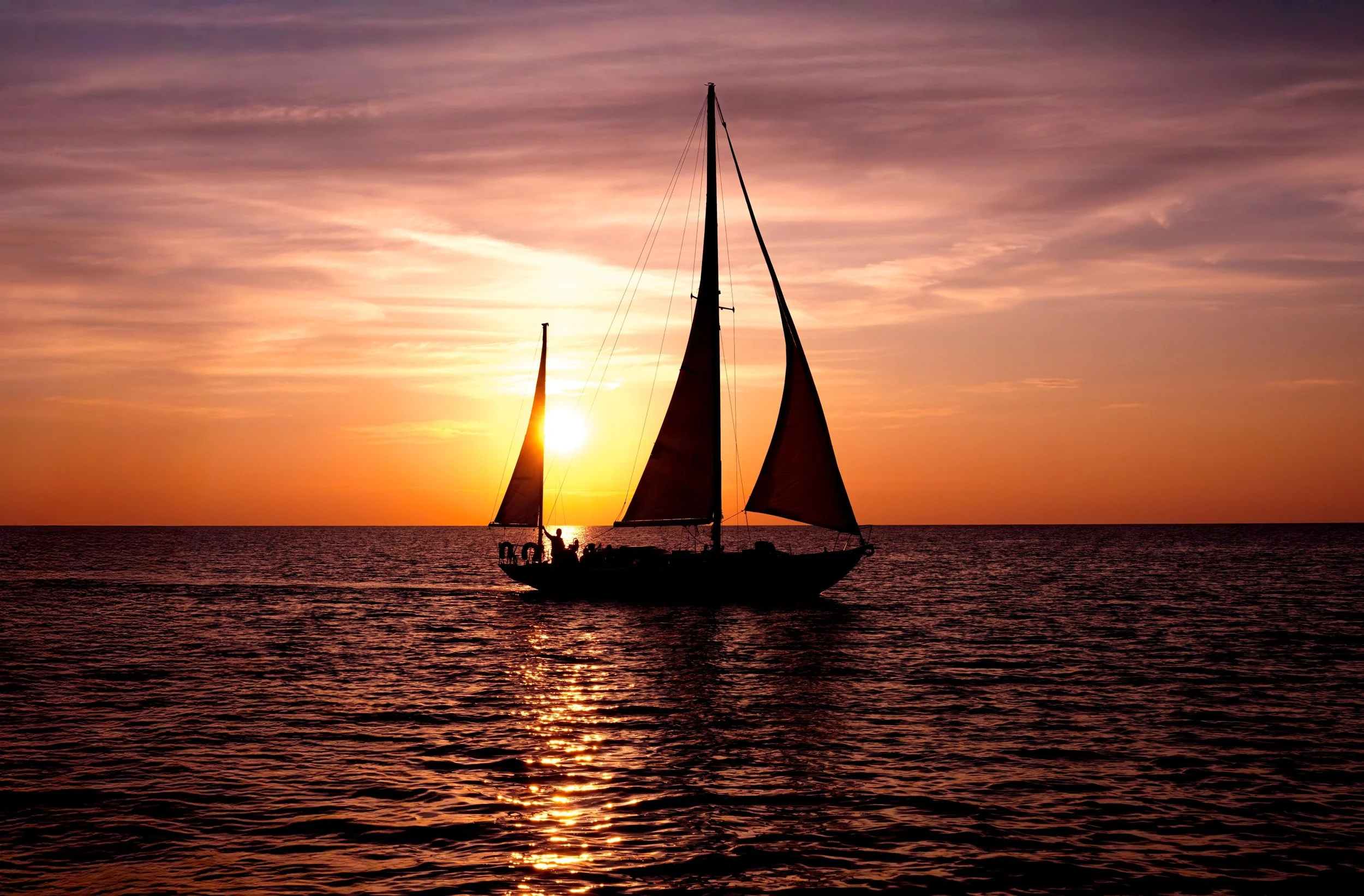 Sailboat sailing on the ocean during sunset with a colorful sky and the sun near the horizon.