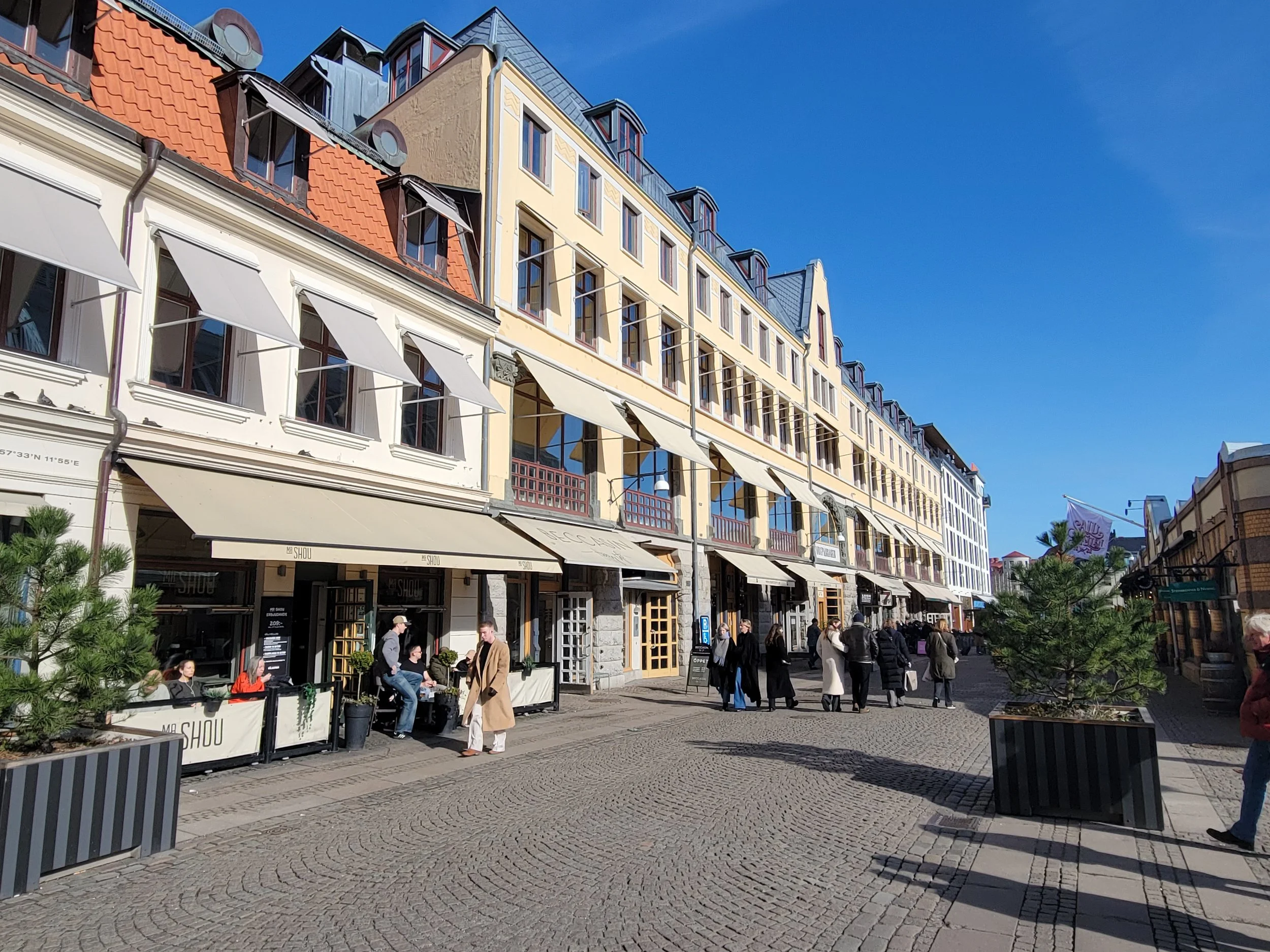 A daytime street scene featuring a row of multi-story buildings with European architectural style, including shops and cafes on the ground floor. People are walking and sitting outside on the cobblestone street, with some wearing winter coats.