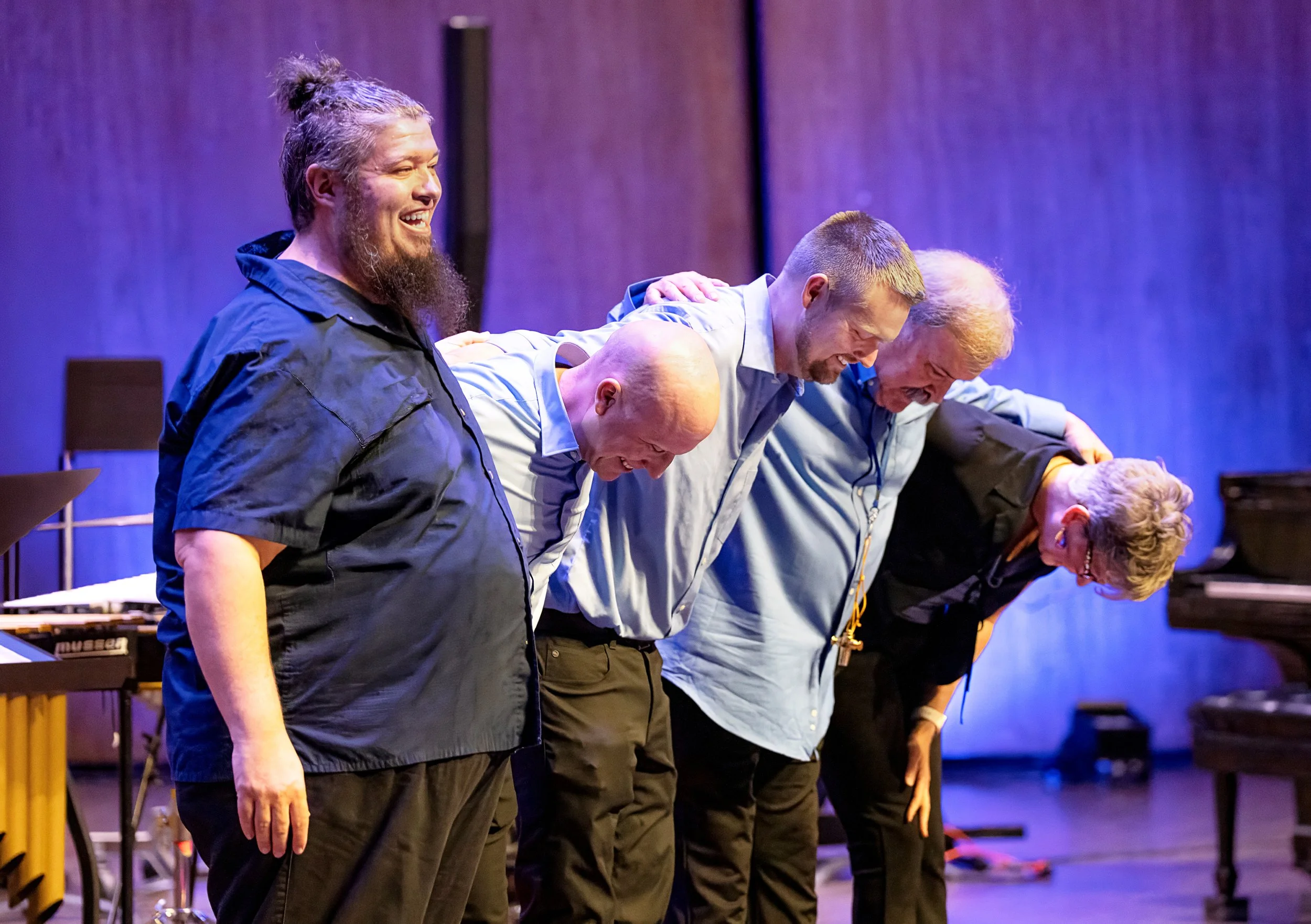 A group of five people standing side by side, bowing their heads and smiling during a performance or event on a stage with purple lighting. They appear to be bowing and acknowledging an audience.