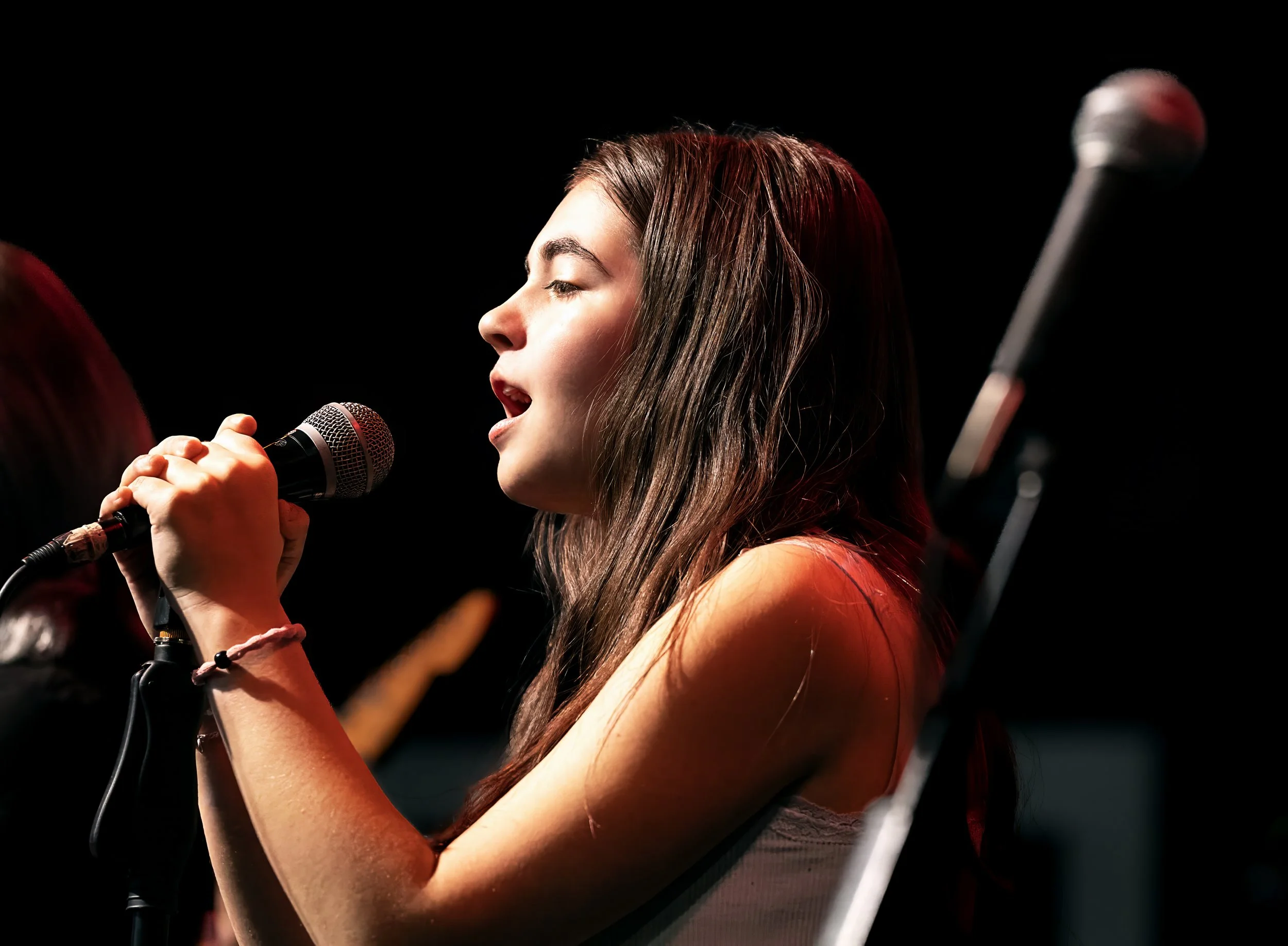 A young woman singing into a microphone during a performance, with dark background and stage lighting.