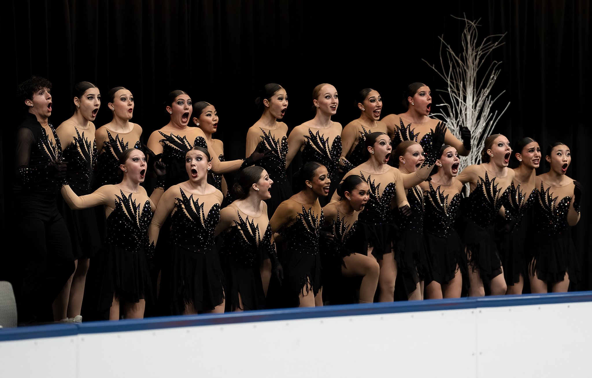 A group of synchronized ice skaters dressed in black and gold costumes, performing on an ice rink against a black background, with some of their mouths open as if singing or cheering.