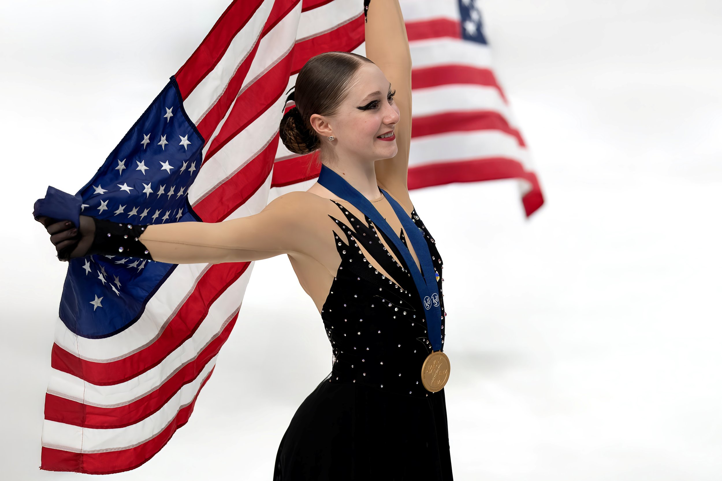 A female ice skater smiling and holding an American flag, wearing a black leotard with rhinestones and a gold medal around her neck, with another American flag in the background.