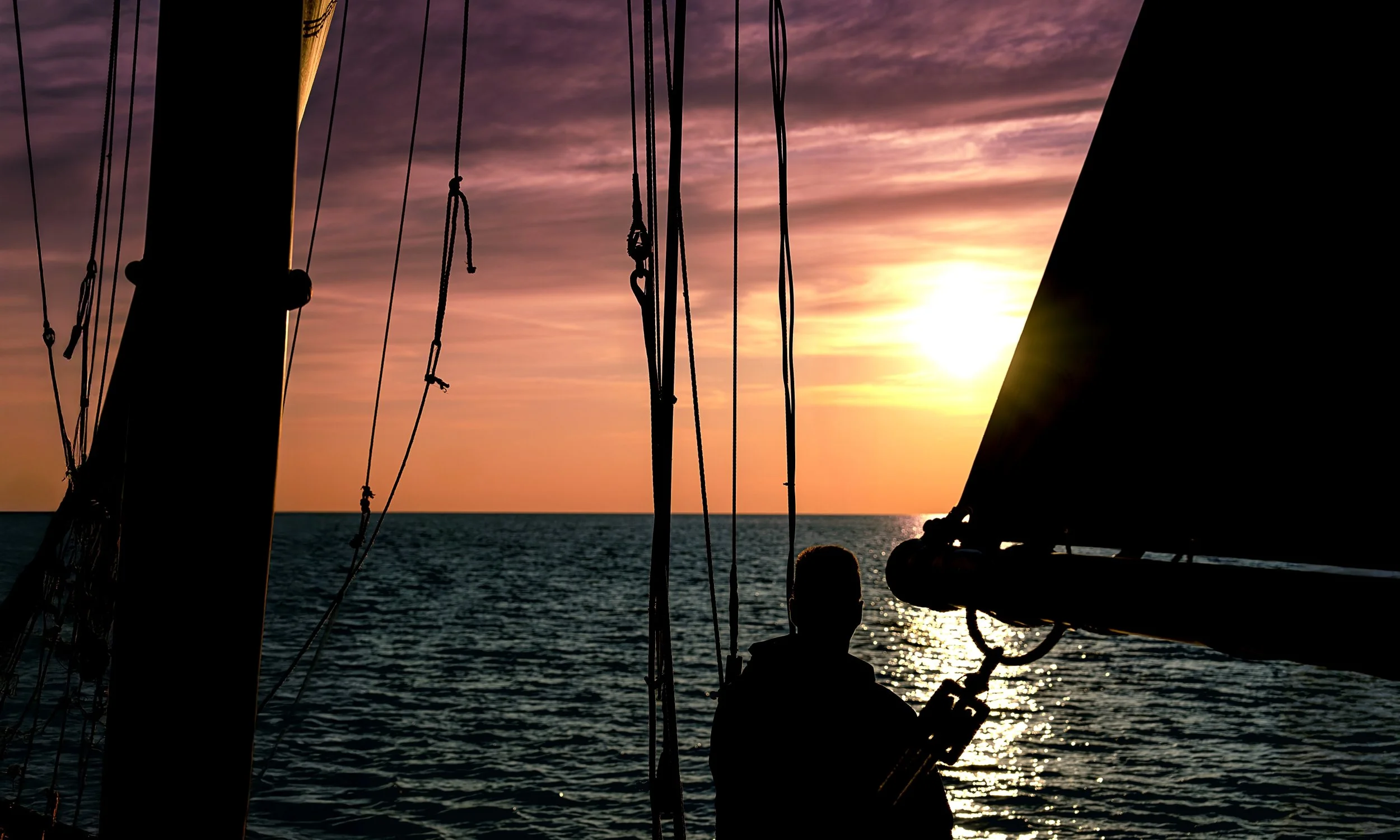 Silhouettes of boat rigging against a sunset over the ocean, with a person sitting on the deck.