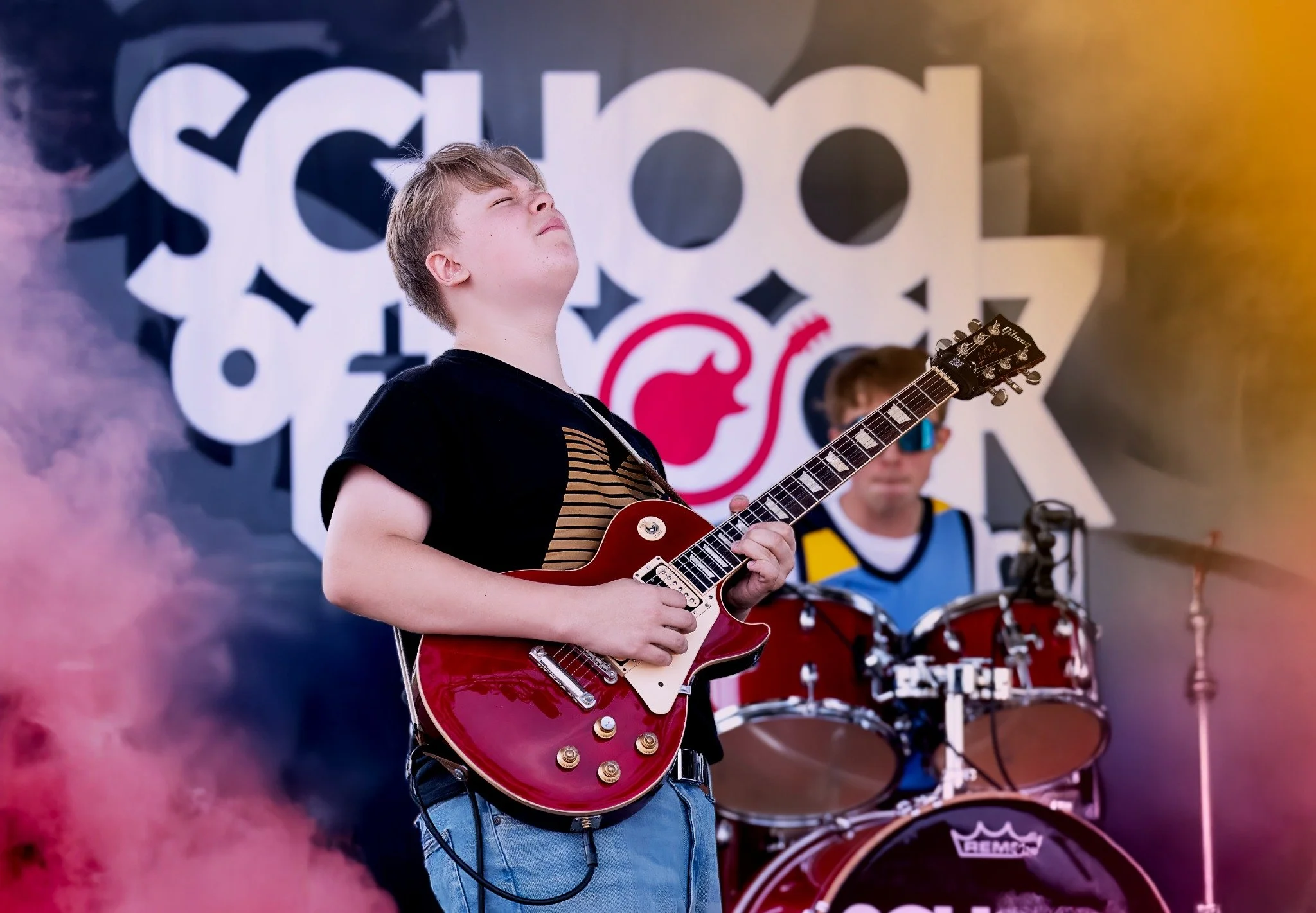A young boy with blonde hair playing a red Gibson electric guitar at a band performance. Another musician is visible in the background playing drums. The stage backdrop features large, abstract white and black text design with a red logo.