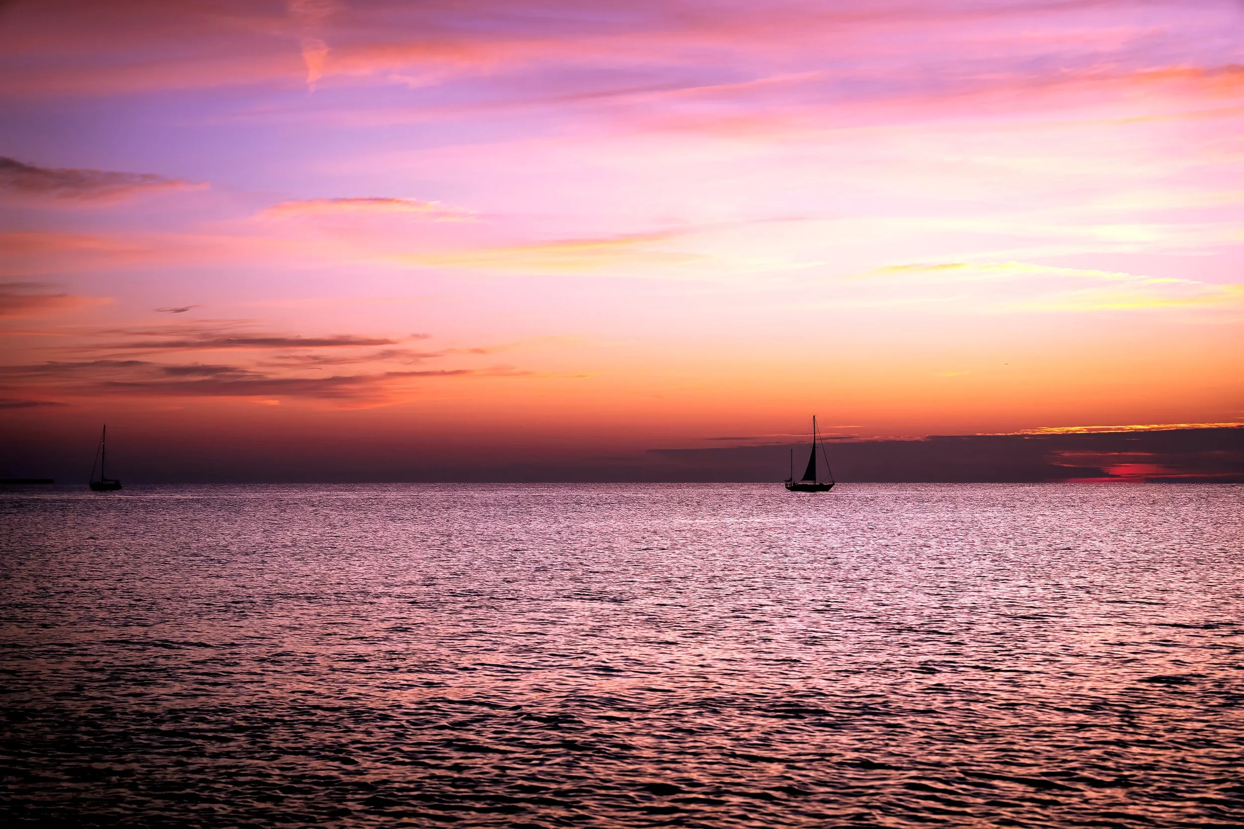 A peaceful ocean sunset with pink, purple, and orange hues in the sky, and silhouettes of sailboats on calm water.