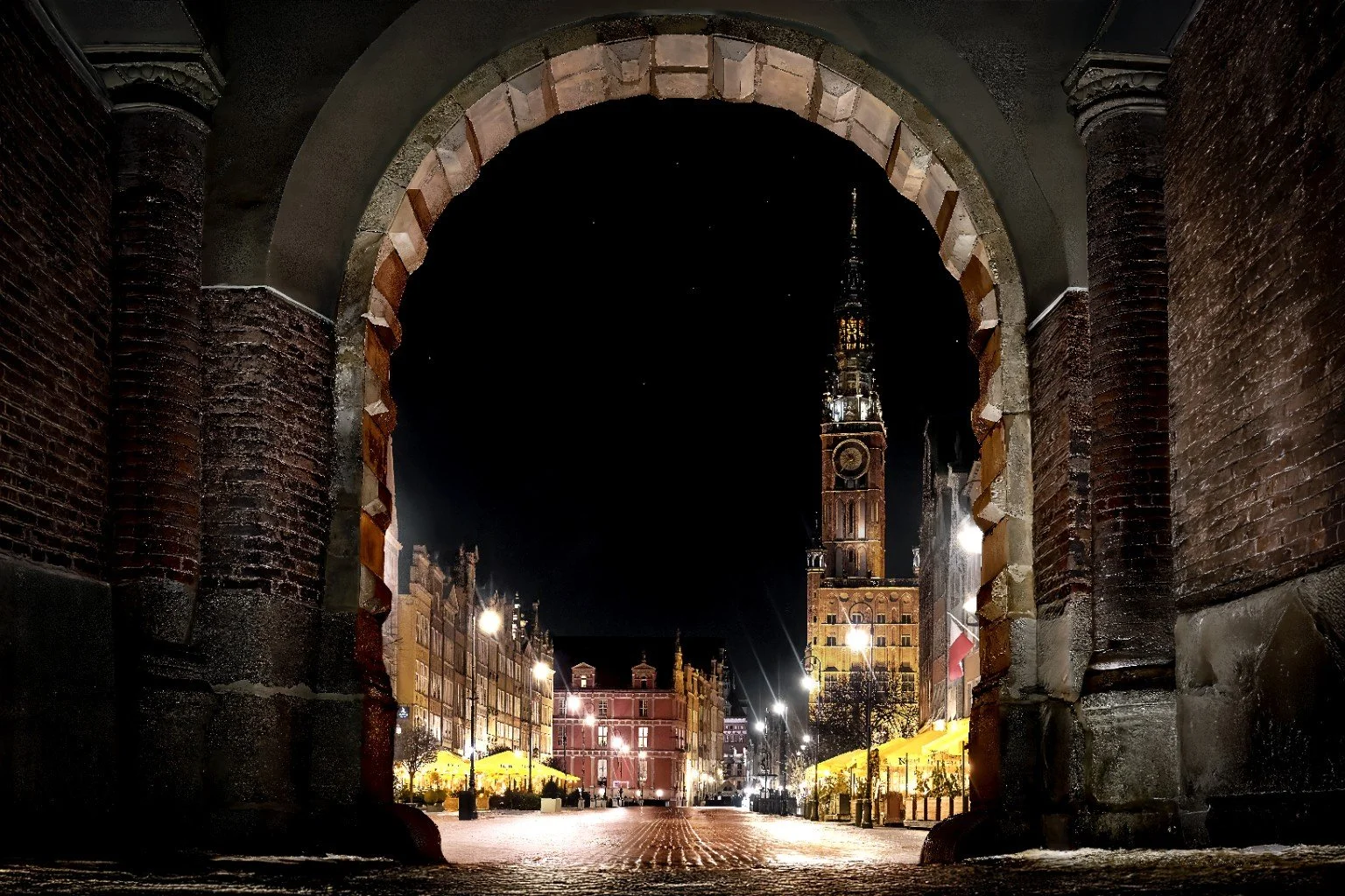 Night view of a historic tower and illuminated buildings through an archway in a European town square.