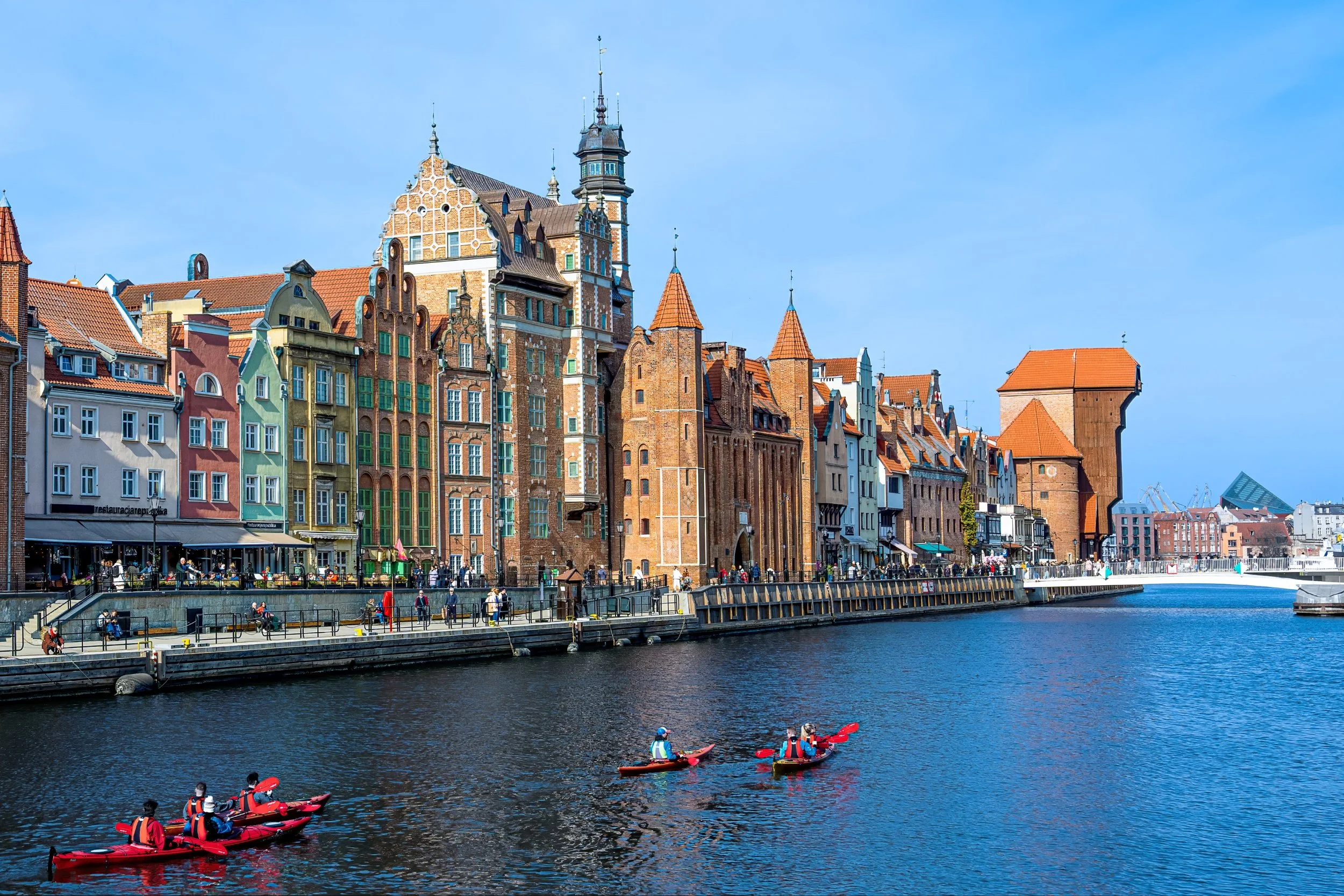 Colorful historic buildings along a riverbank with people kayaking on the water and people walking along the promenade.