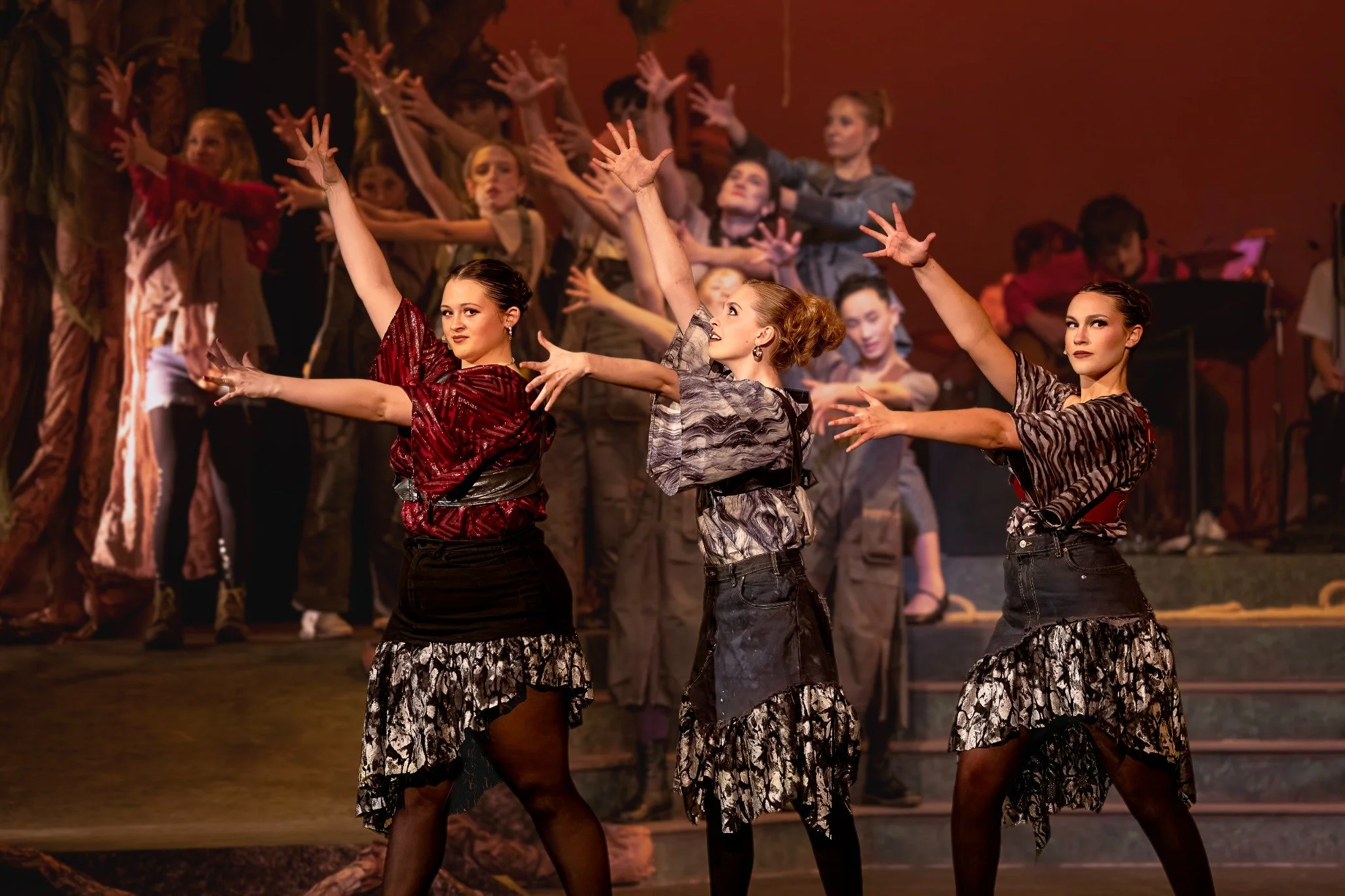 Three women in vintage-style costumes performing a dance on stage, with a group of children in similar costumes in the background.
