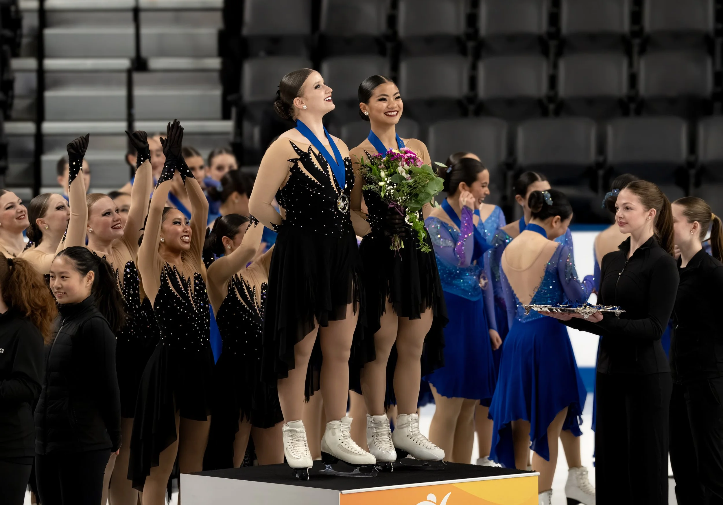 Two female figure skaters standing on a podium, smiling with medals around their necks and holding flowers, after a competition. They are surrounded by other skaters clapping and celebrating.