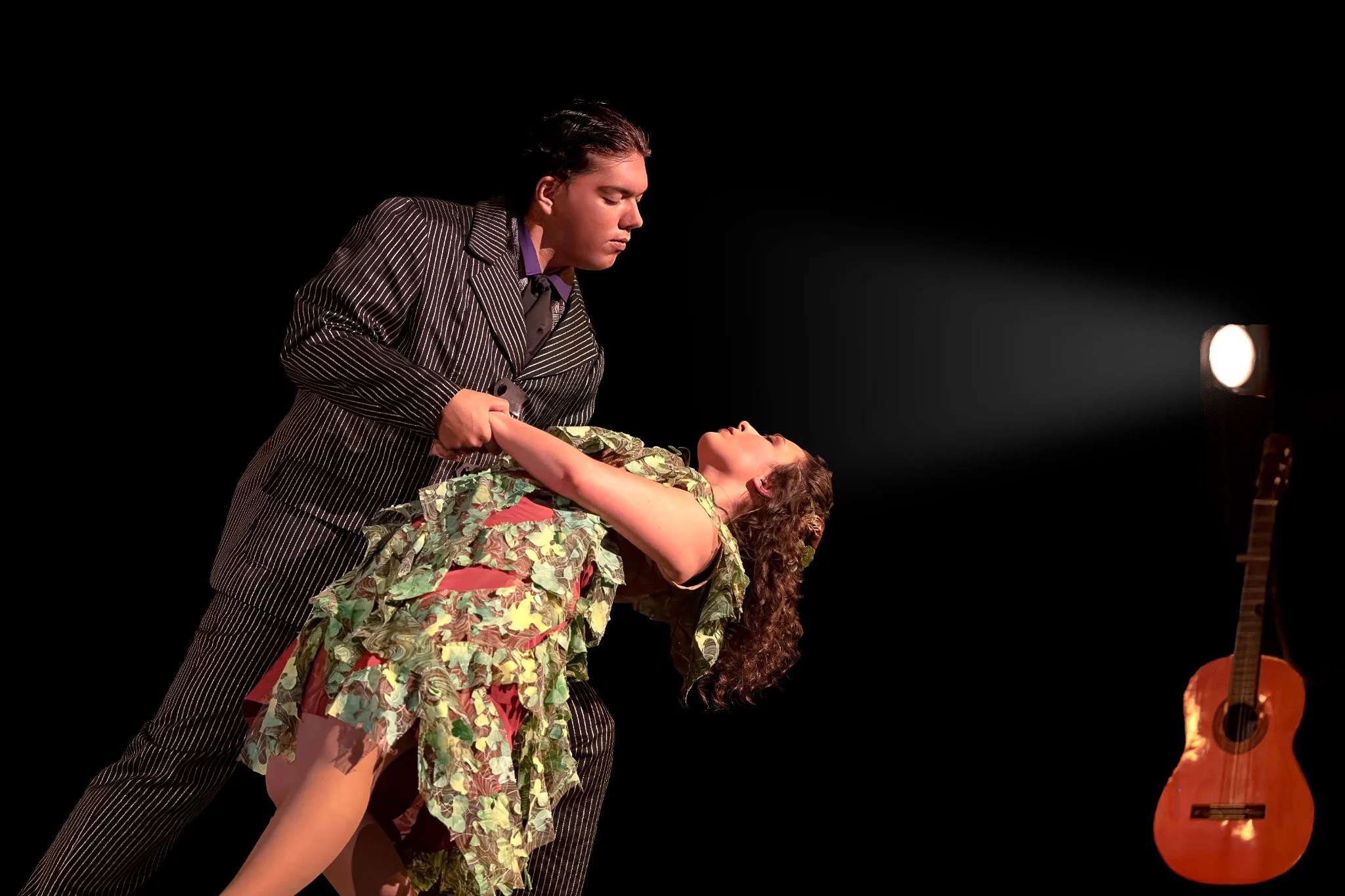 A man in a pinstripe suit holds and looks at a woman who is leaning back in a floral dress, on a dark stage with a spotlight and a guitar in the background.