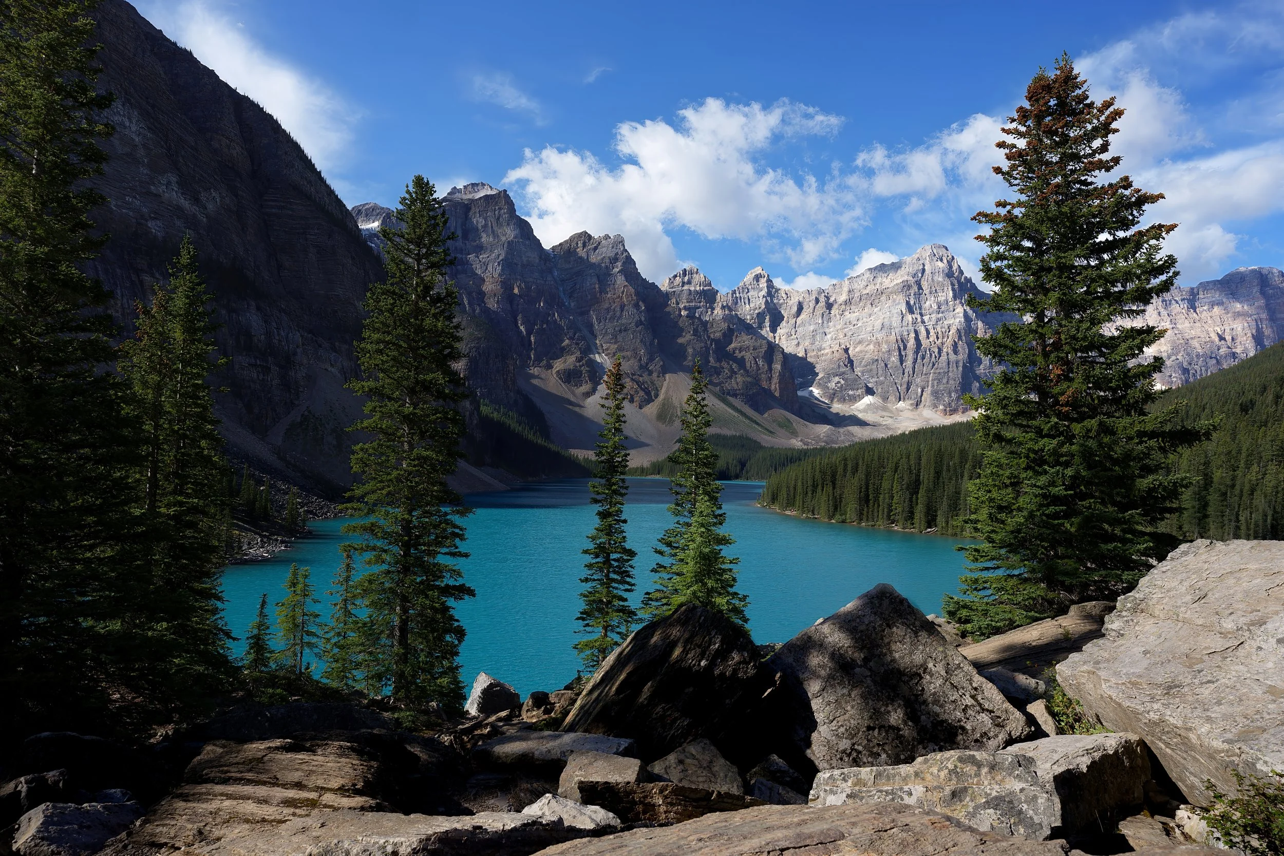 A scenic view of a turquoise lake surrounded by dense evergreen trees and rugged mountains under a partly cloudy blue sky.