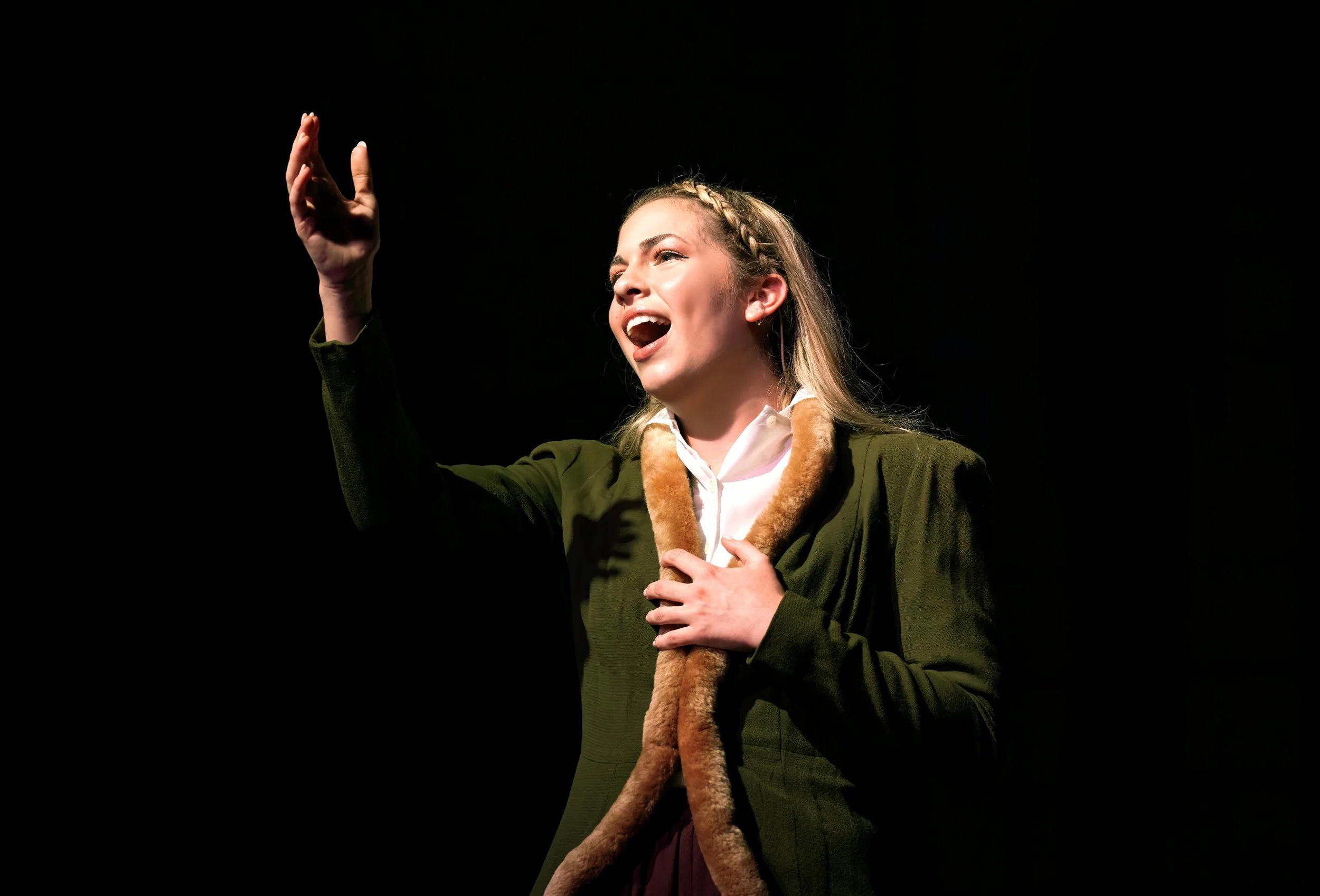 A young woman with braided hair singing or speaking passionately on stage, holding a fur-lined scarf and raising her hand.
