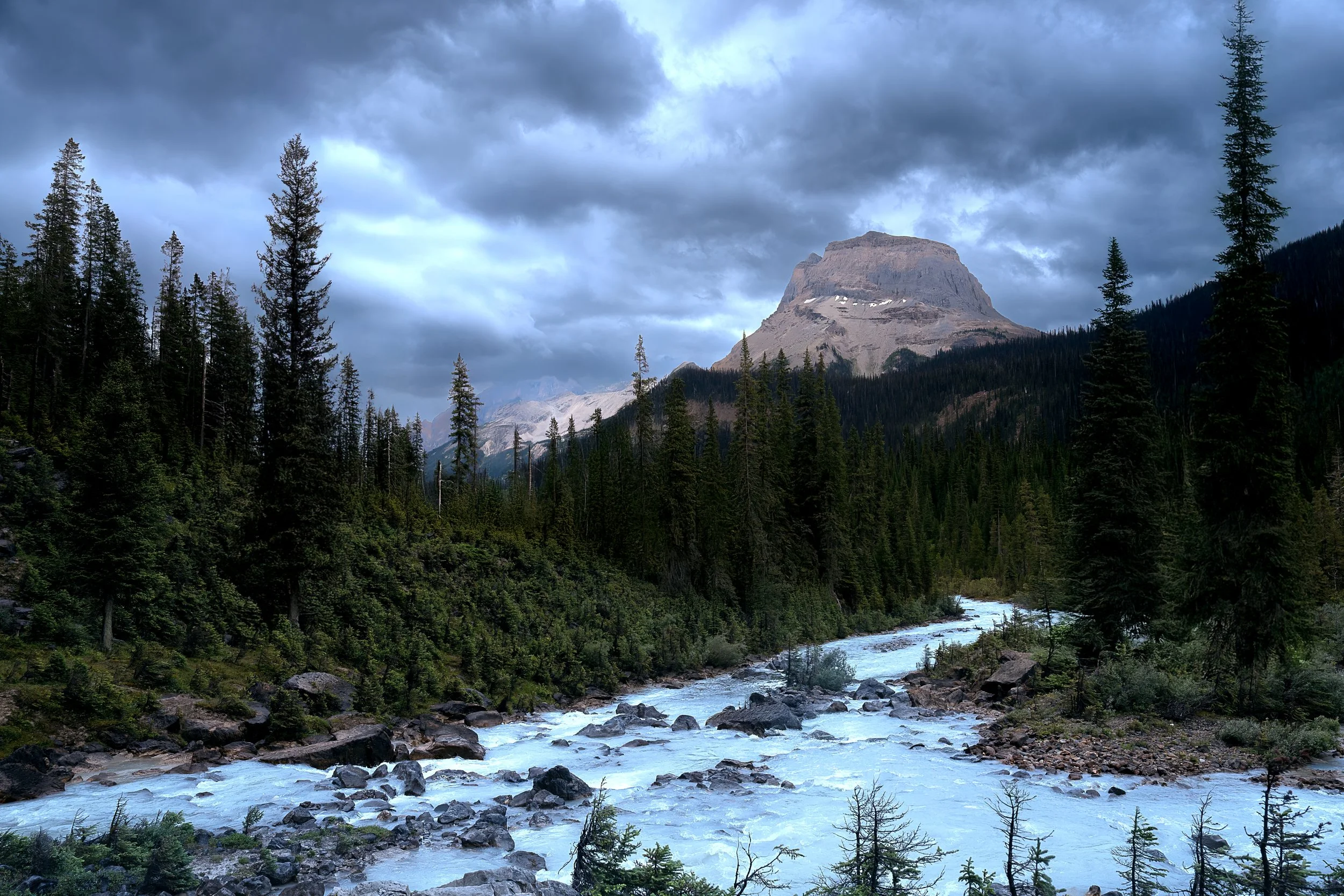 A mountain surrounded by a forest with a river flowing in the foreground and cloudy skies overhead.