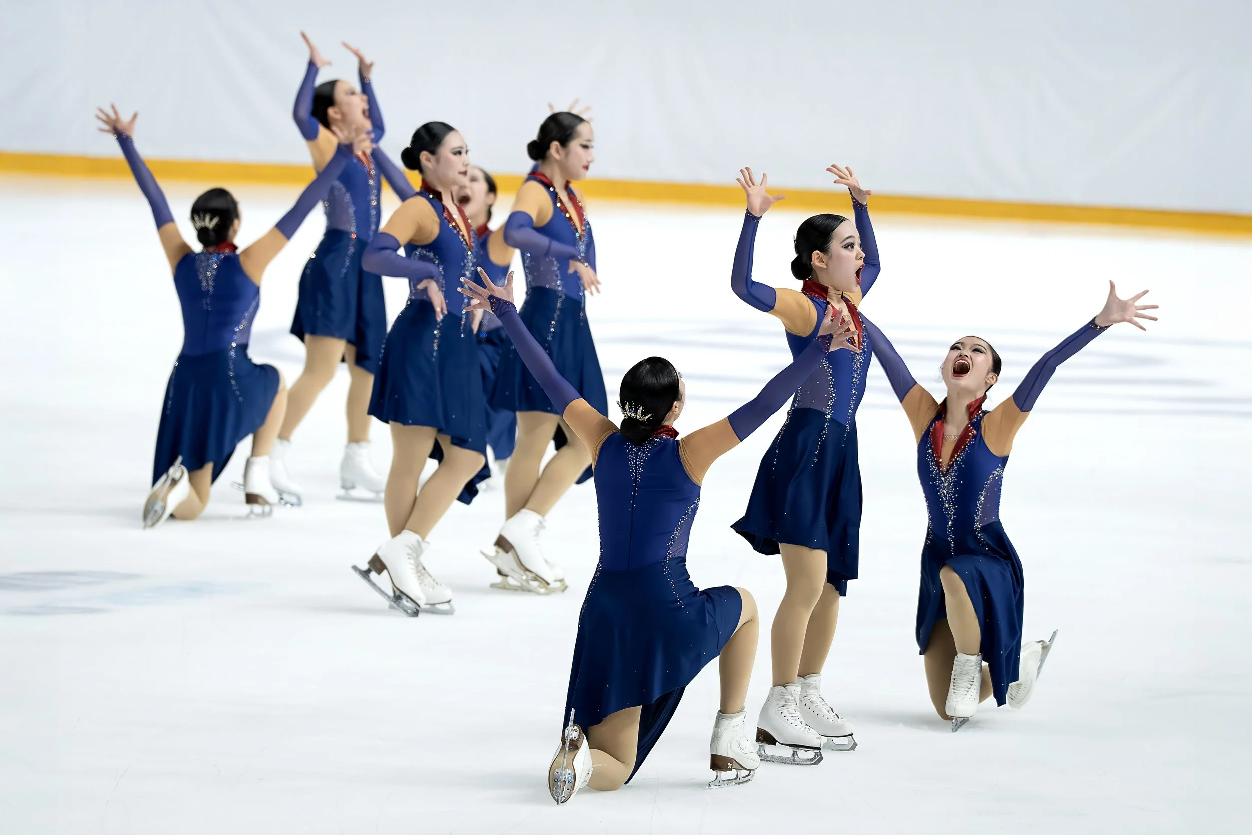 A group of young female figure skaters in matching blue and gold costumes performing a choreographed routine on the ice rink, with some kneeling and others standing with arms raised and wide open mouths.
