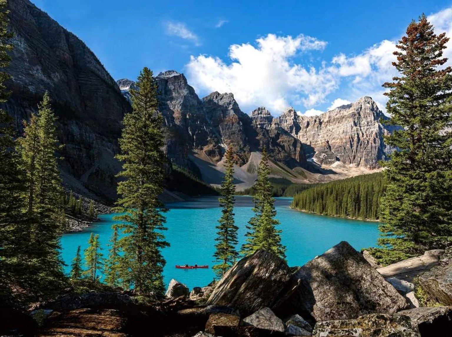 Scenic view of a turquoise lake surrounded by tall pine trees and mountains with a partly cloudy sky.