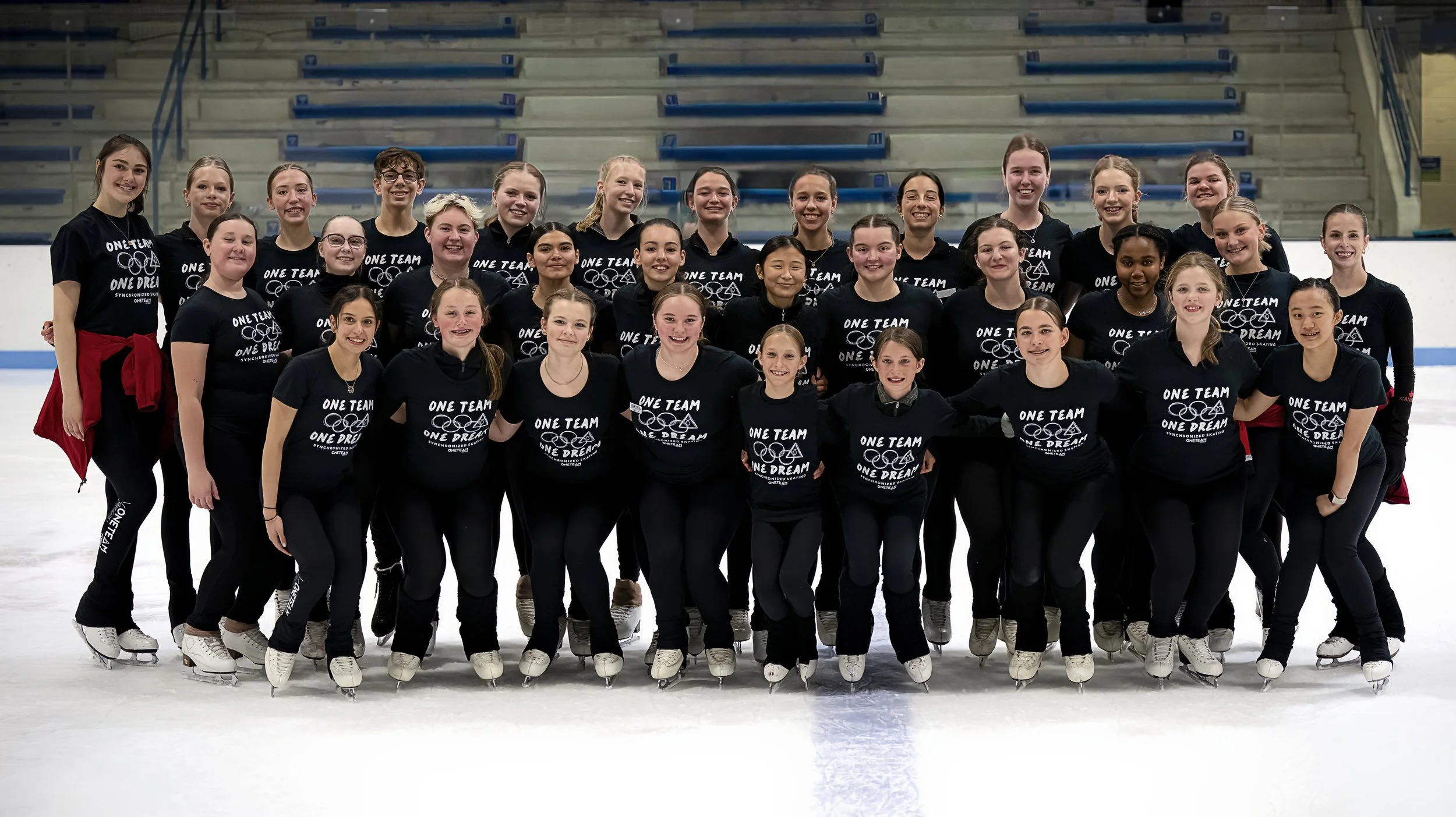 Group of young female figure skaters standing on an ice rink, smiling at the camera, wearing matching black shirts with 'One Team, One Dream' written on them.