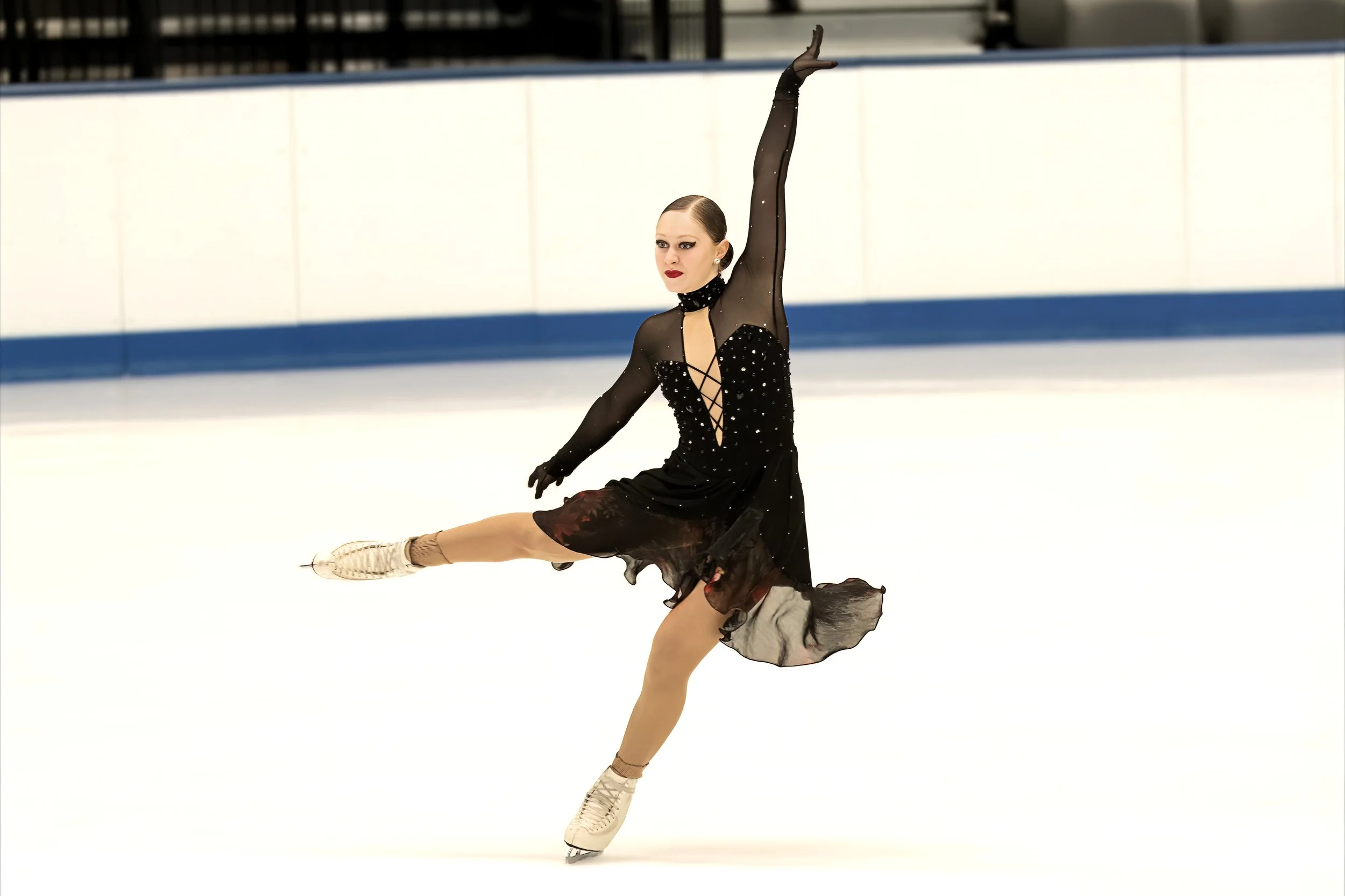 An ice skater wearing a black dress and figure skates performing a move on the ice rink.