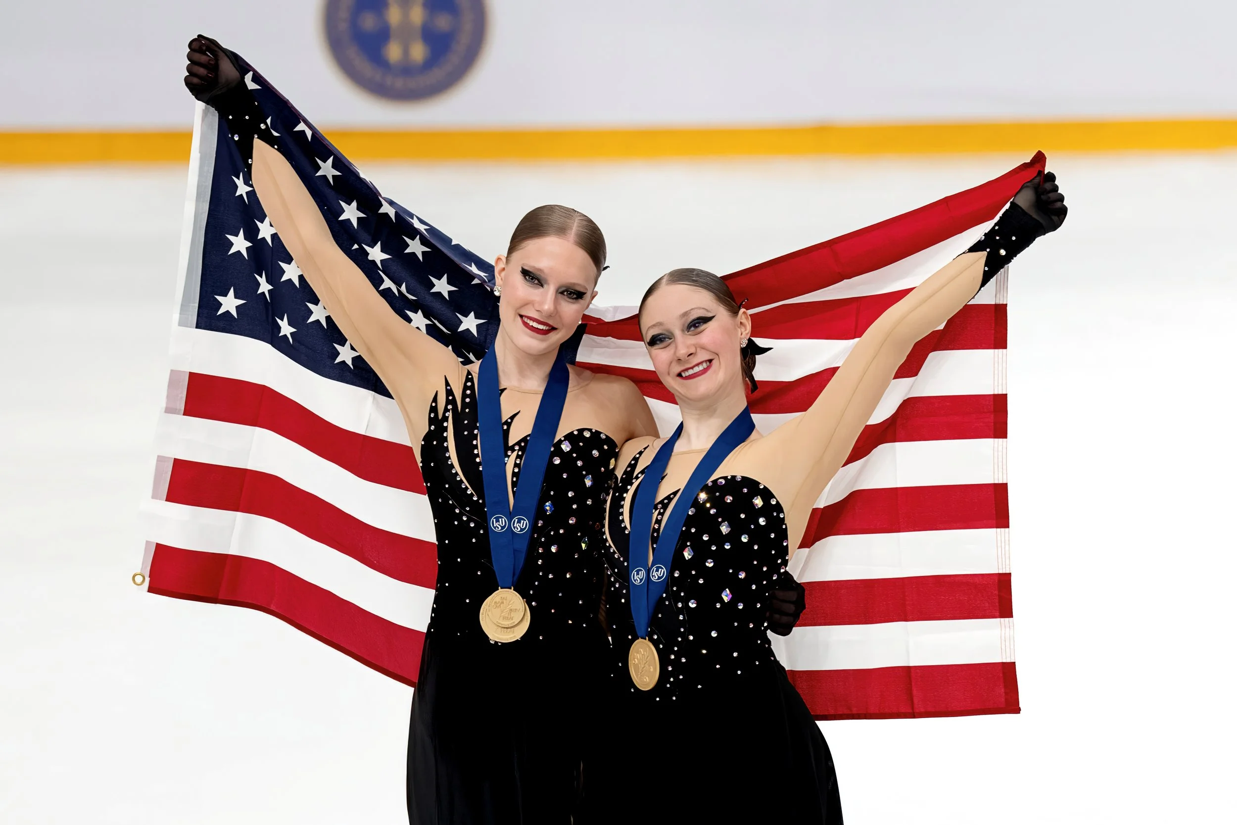 Two female figure skaters wearing black costumes with rhinestones, holding an American flag, and wearing medals around their necks after a competition.