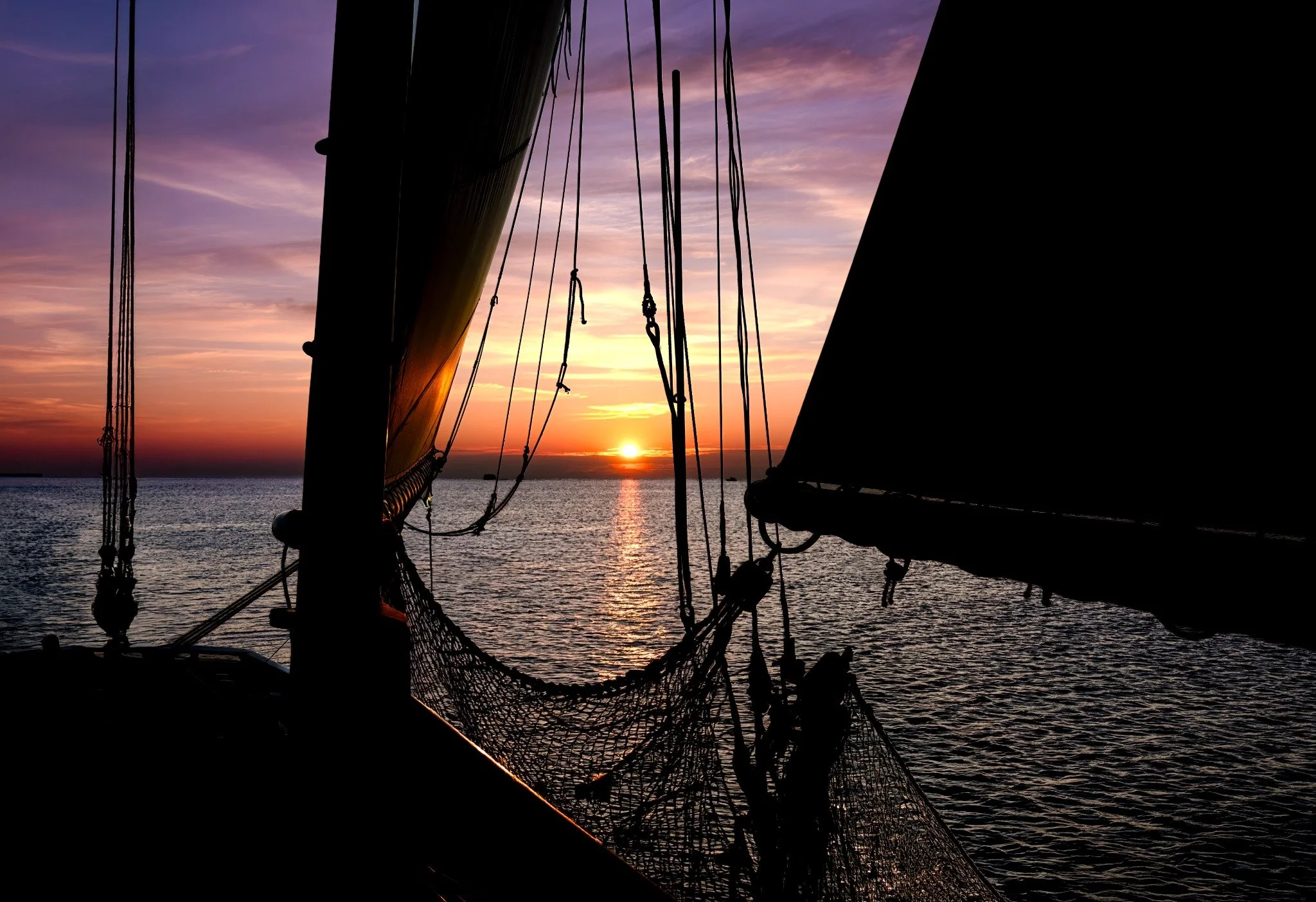 Silhouette of a boat's mast, rigging, and netting against a colorful sunset over the ocean