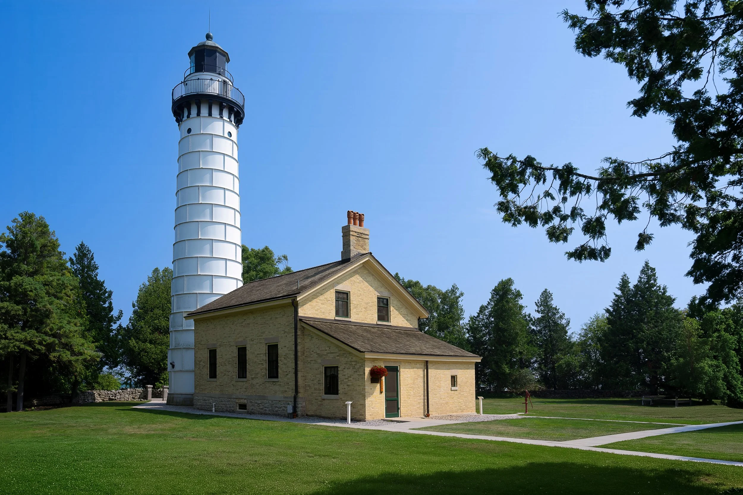 A historic lighthouse with a white, cylindrical structure topped with a dark lantern room, situated behind a two-story yellow brick house with a chimney, green lawn, surrounded by trees under a clear blue sky.