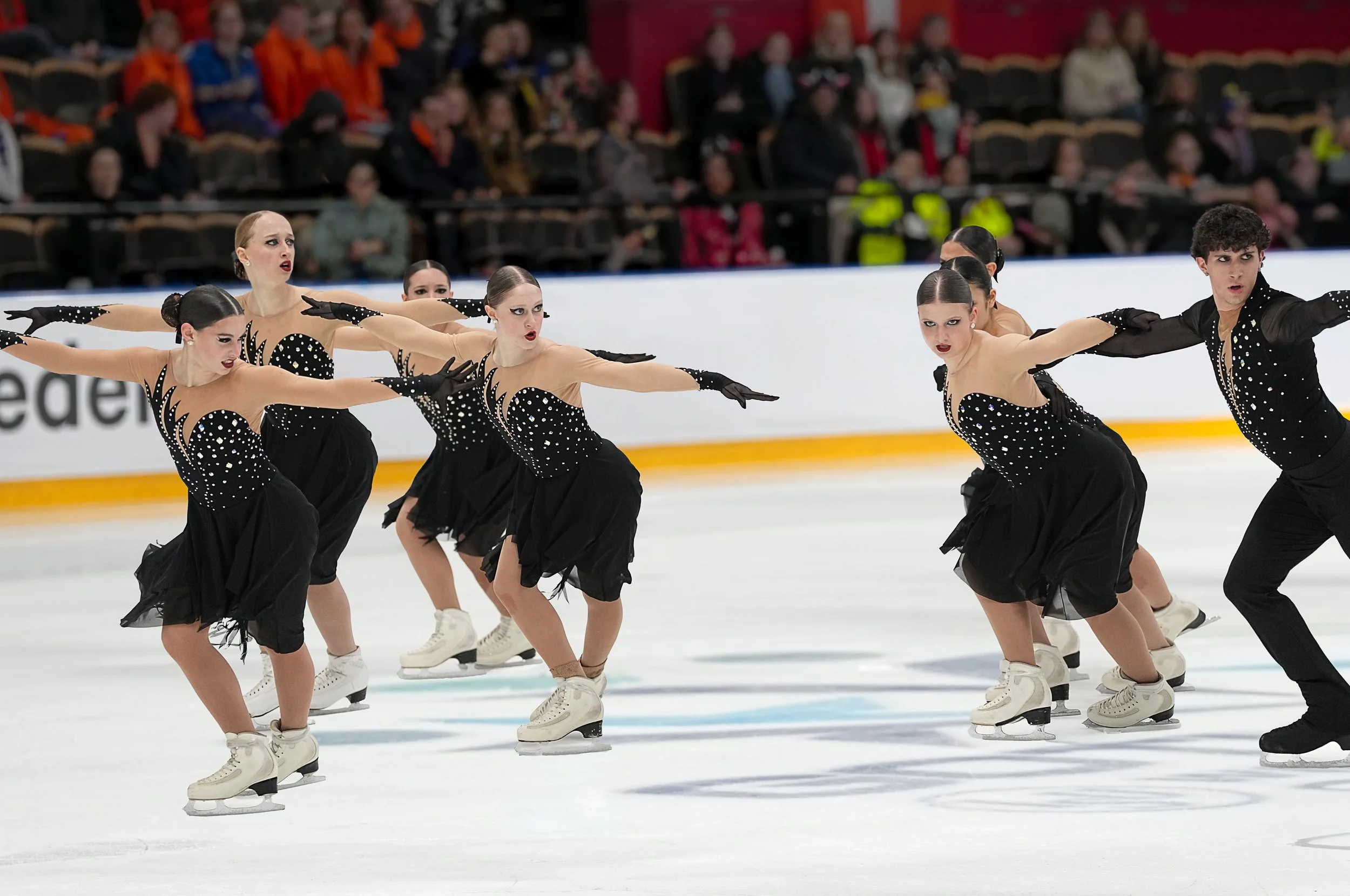 A group of figure skaters on ice performing a synchronized routine wearing black and beige costumes with rhinestones, and white figure skates.